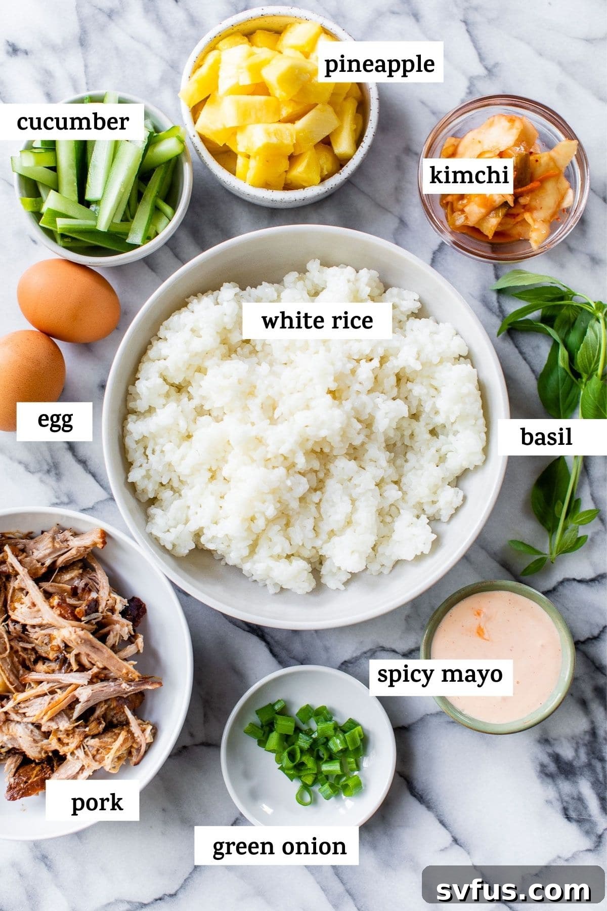 A flat lay image displaying all the fresh ingredients needed to make Hawaiian Bibimbap, including rice, pulled pork, pineapple, cucumber, kimchi, basil, and spicy mayo