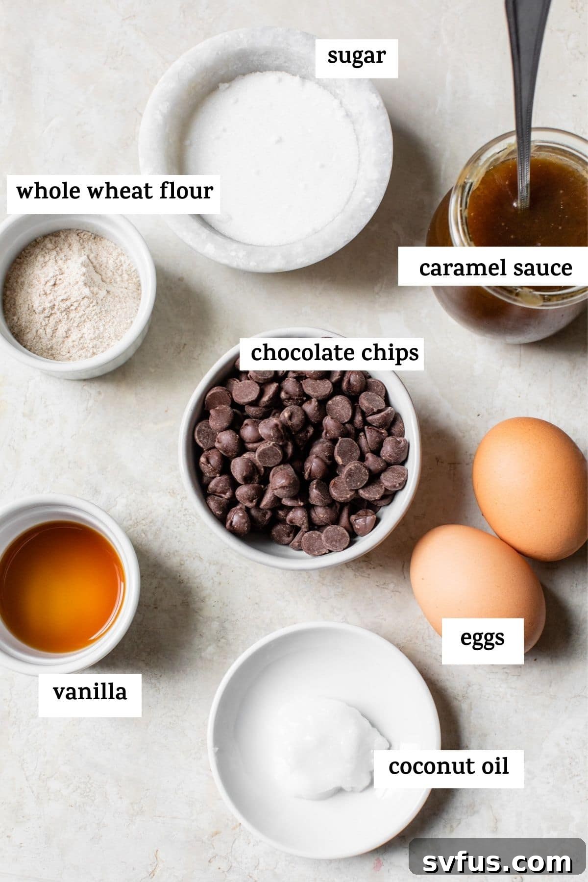 Various baking ingredients for chocolate caramel lava cake laid out on a surface, including chocolate chips, coconut oil, eggs, flour, sugar, and a jar of caramel sauce.