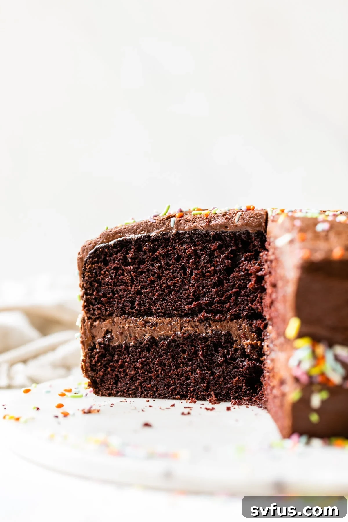 A slice of rich chocolate vegan birthday cake on a white plate, topped with creamy dairy-free chocolate buttercream icing and colorful sprinkles.