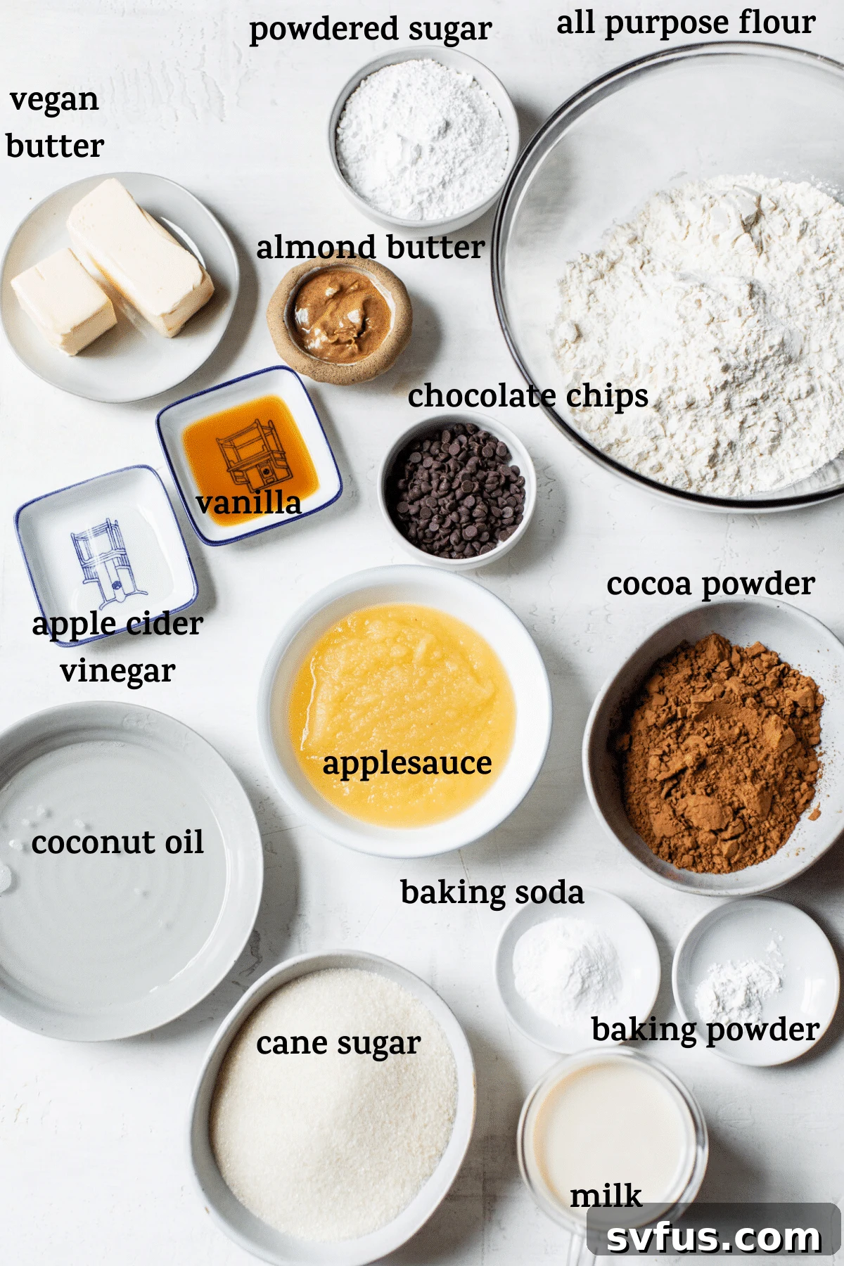 A collection of cake ingredients laid out on a surface, including flour, sugar, cocoa powder, milk, applesauce, and chocolate chips.