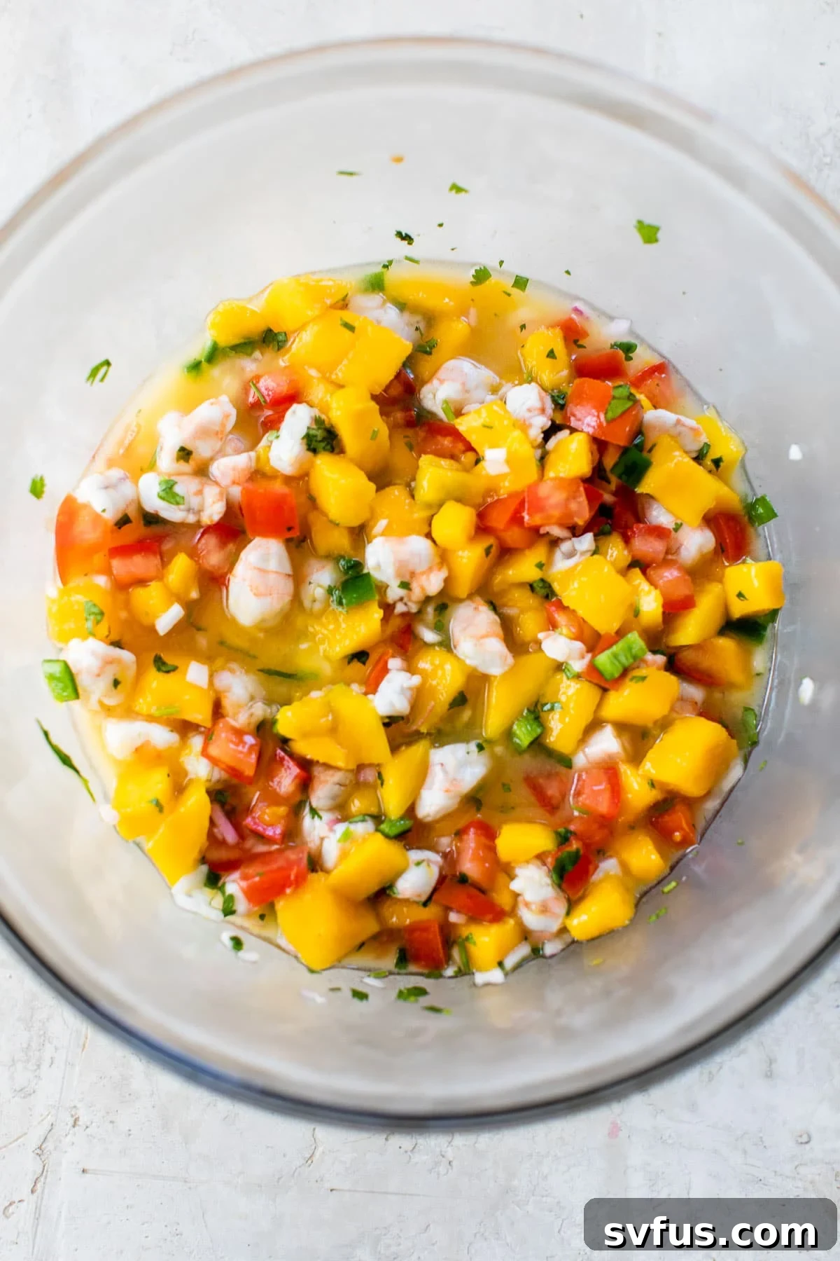 A large, clear glass bowl brimming with the colorful mango shrimp ceviche, showcasing the fresh mixture of pink shrimp, orange mango, and red tomatoes.