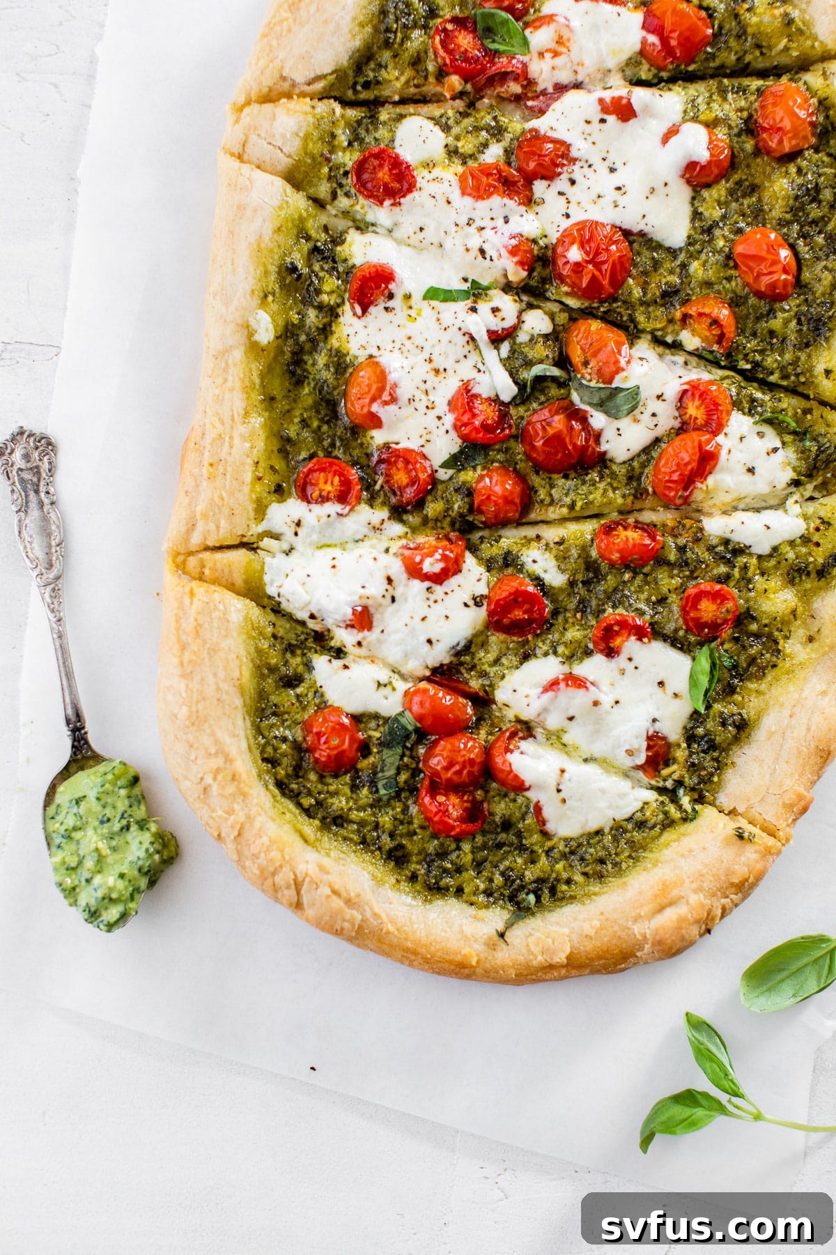 Sliced Kale Pesto Pizza on parchment paper, showcasing its vibrant green pesto, red tomatoes, and melted burrata.