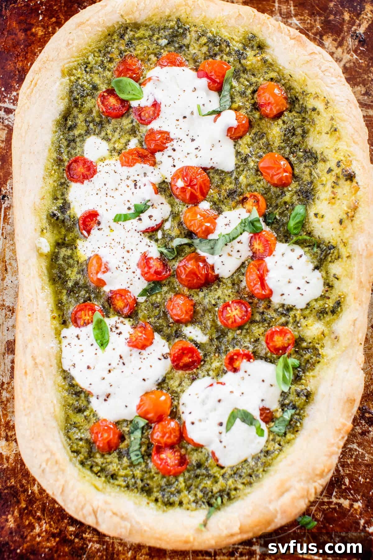 Freshly baked Kale Pesto Pizza on a baking sheet, with melted burrata and roasted tomatoes, garnished with fresh basil.