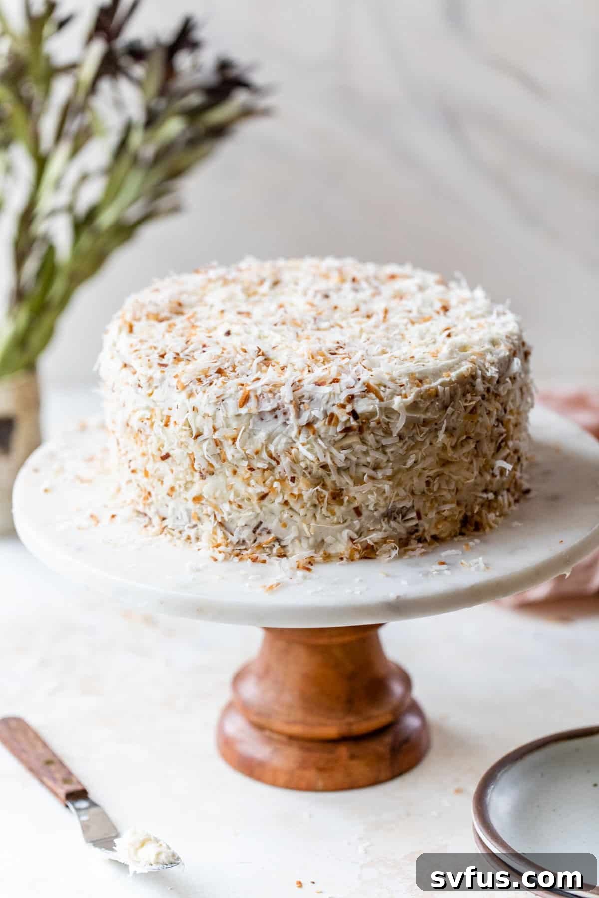 A stunning vegan coconut cake on a cake stand, elegantly decorated with toasted shredded coconut on top and around the sides.