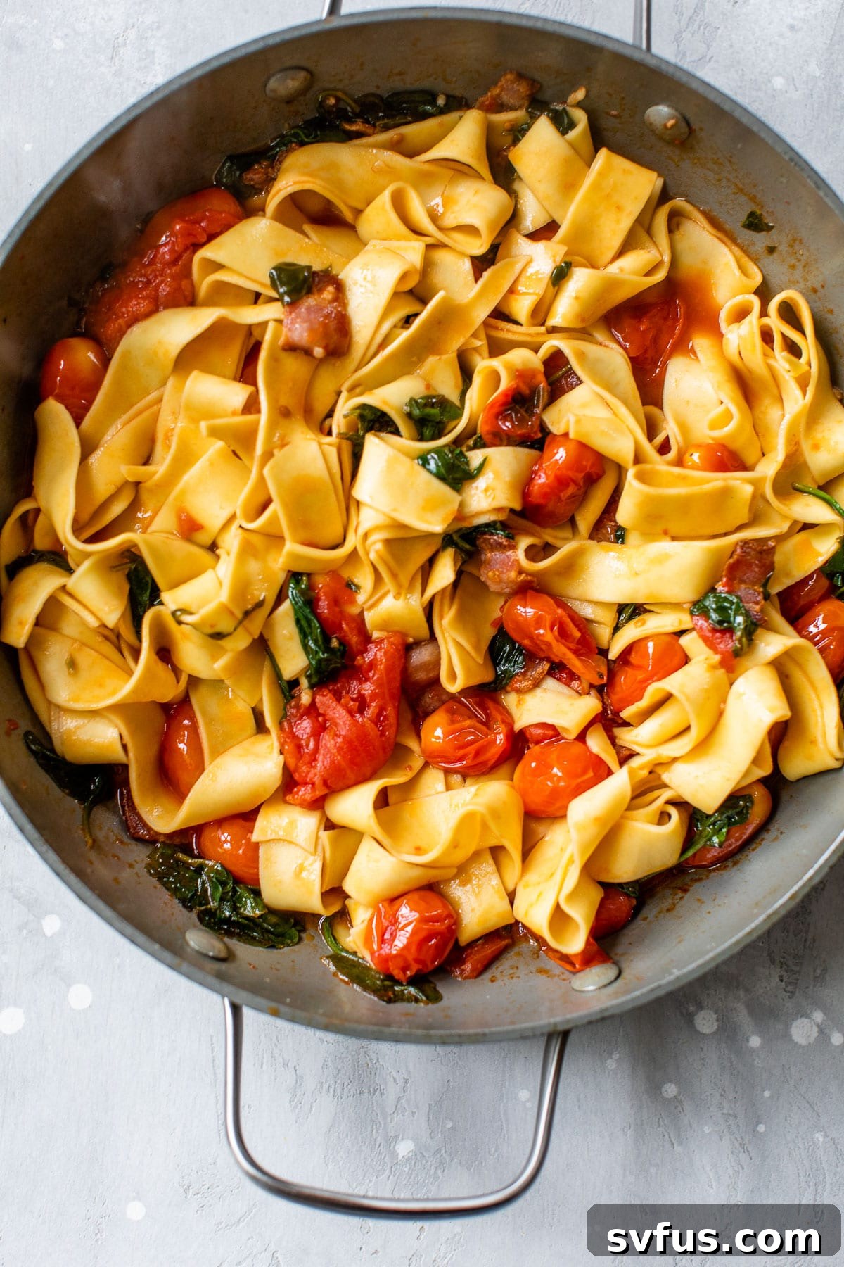 A large gray skillet filled with perfectly cooked pasta noodles, vibrant grape tomatoes, and wilted spinach, simmering in a flavorful sauce.
