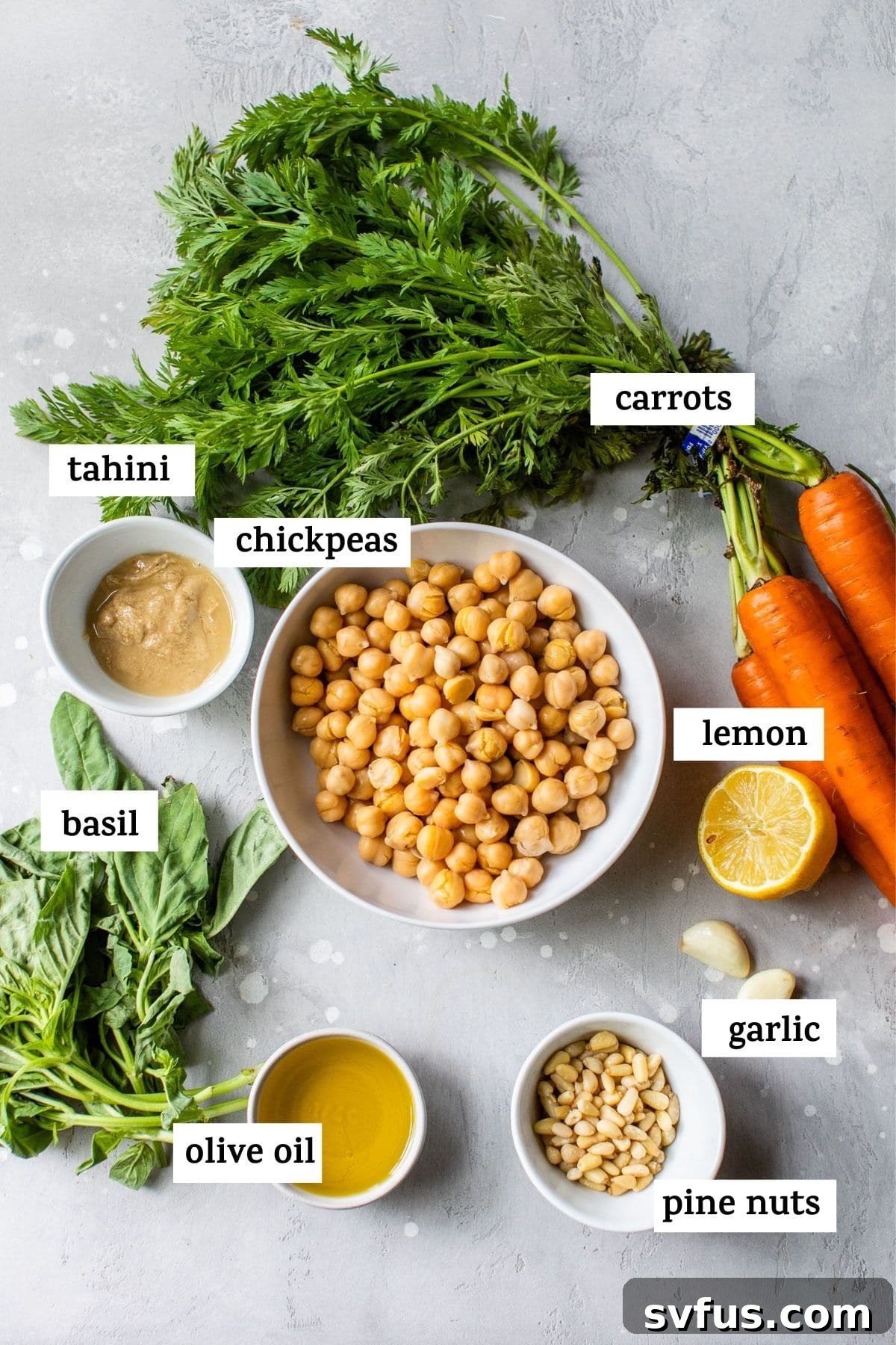 An overhead shot of fresh ingredients laid out on a white surface, including chickpeas, garlic, lemon, tahini, olive oil, basil, pine nuts, and carrot tops, with text overlay 'Hummus Ingredients'.