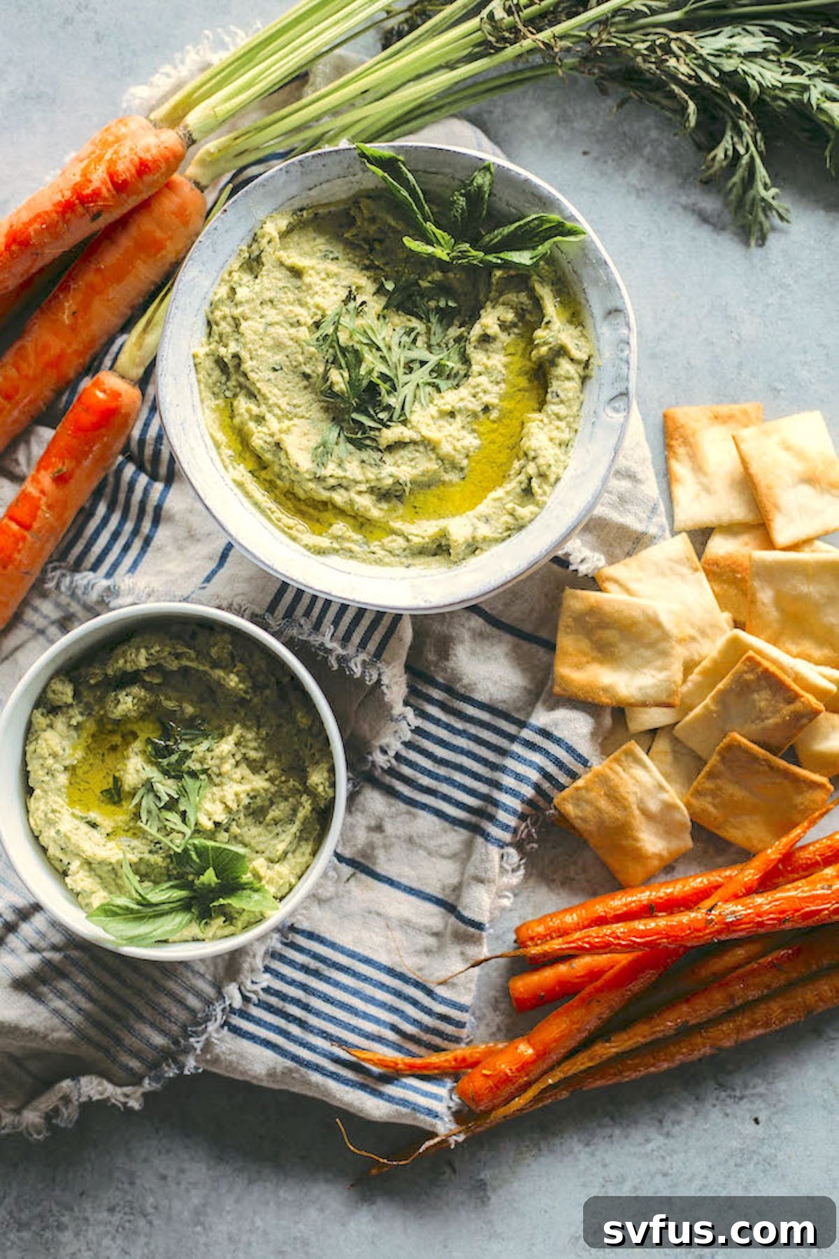 A beautifully arranged table featuring a bowl of vibrant green pesto hummus, surrounded by roasted carrot sticks and crispy pita chips for dipping, creating an inviting spread.