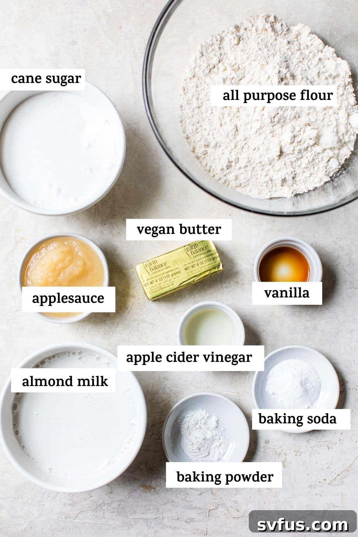 A top-down view of essential ingredients neatly arranged for baking a vegan vanilla cake, including flours, sugars, and liquid components.
