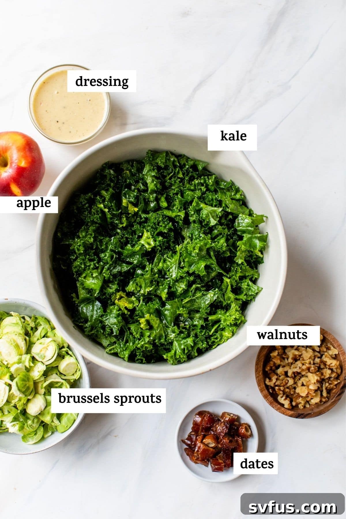 A vibrant flat lay of fresh salad ingredients for a Kale, Apple, and Brussels Sprouts Salad, featuring a pile of green kale, crisp red apples, raw Brussels sprouts, and dried dates, ready for preparation.