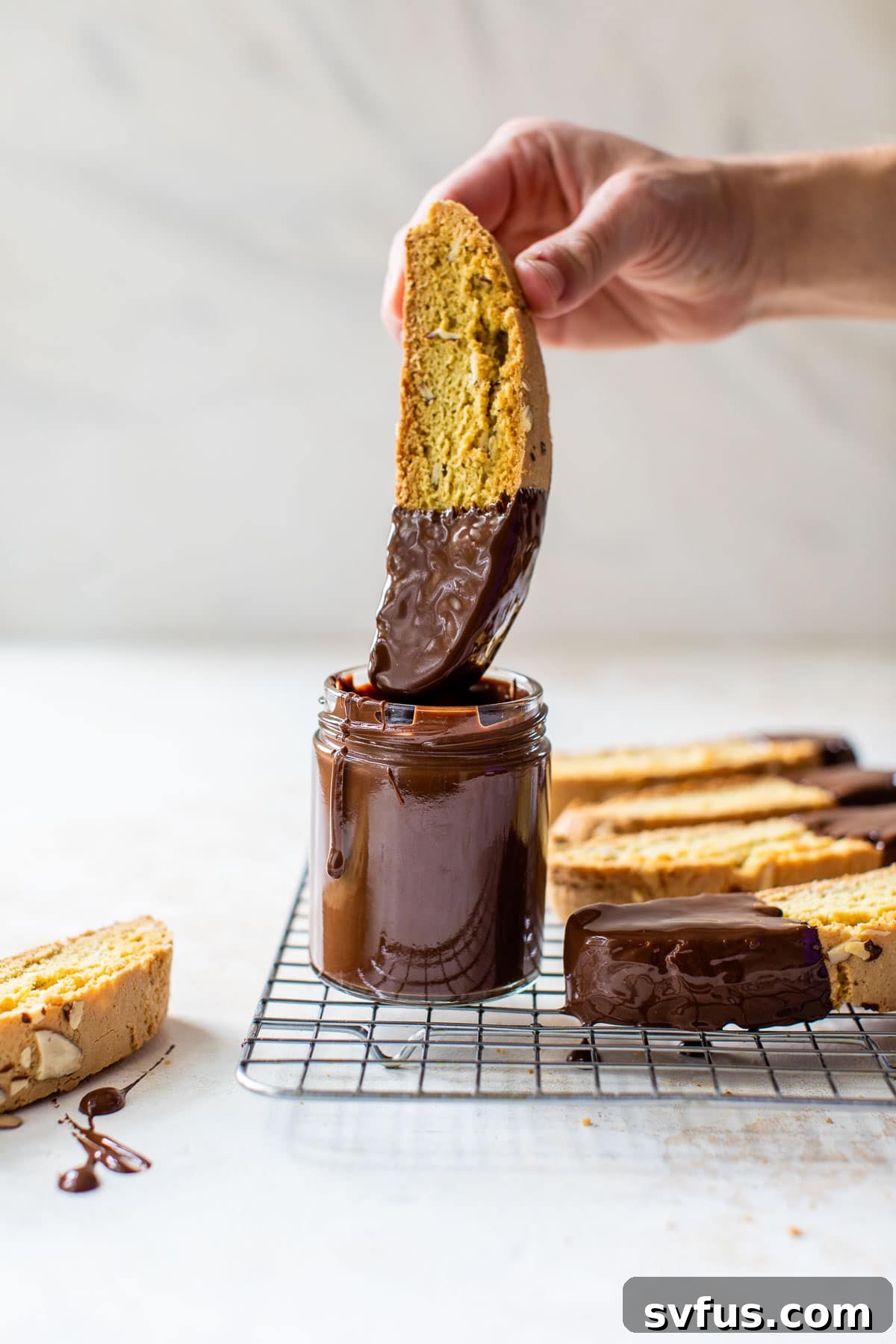 A hand dipping a piece of freshly baked almond biscotti into a jar of melted dark chocolate, with other dipped biscotti on a cooling rack.