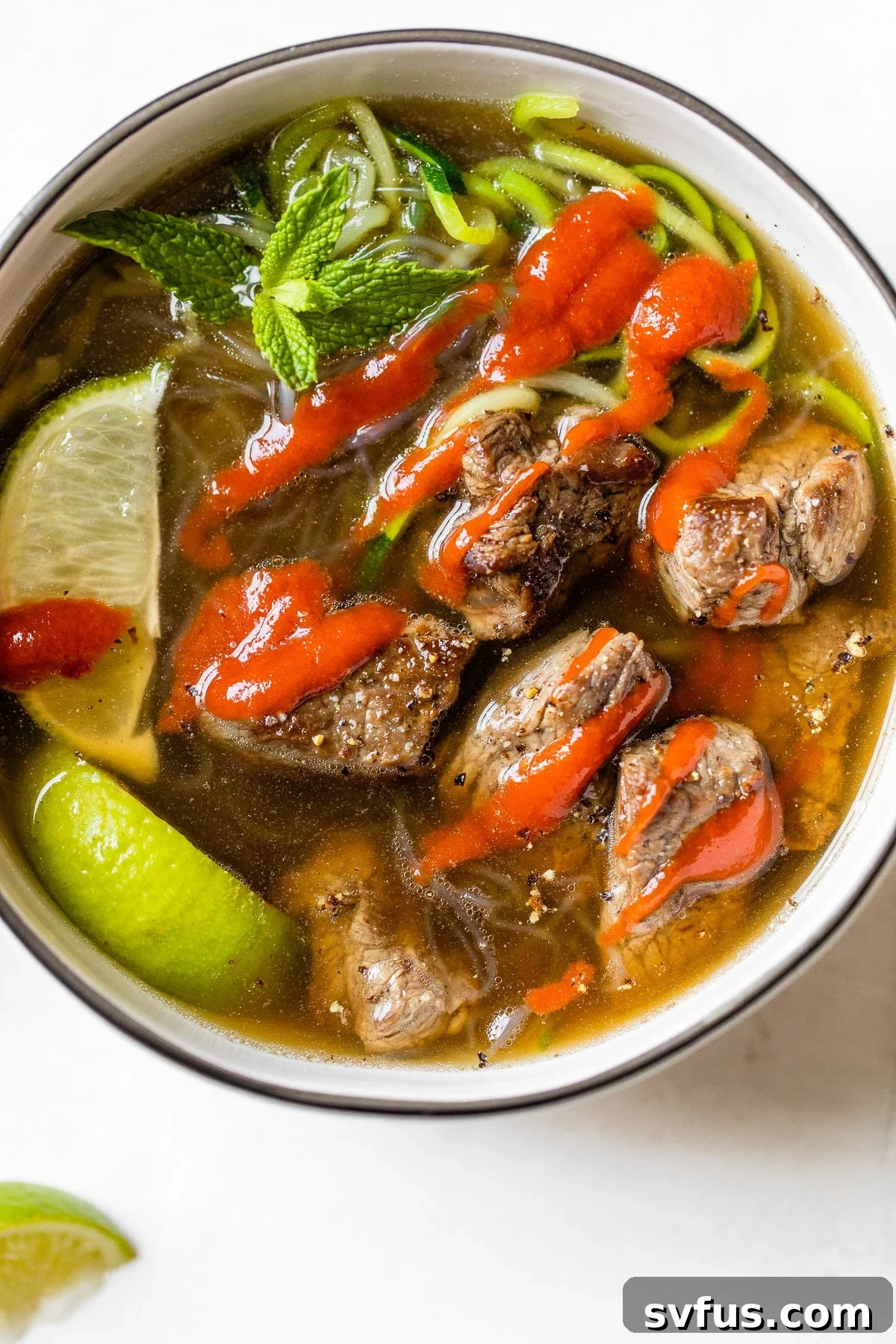 A steaming bowl of Paleo Beef Pho with tender beef, zucchini and kelp noodles, garnished with fresh herbs and a drizzle of sriracha.