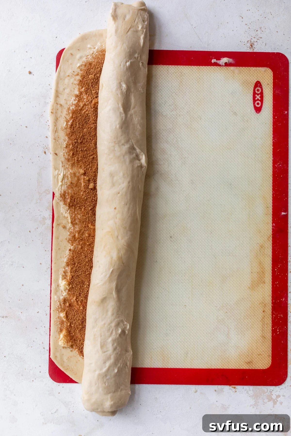 Overhead view of a half-rolled log of vegan cinnamon roll dough on a baking mat, showing the filling inside.