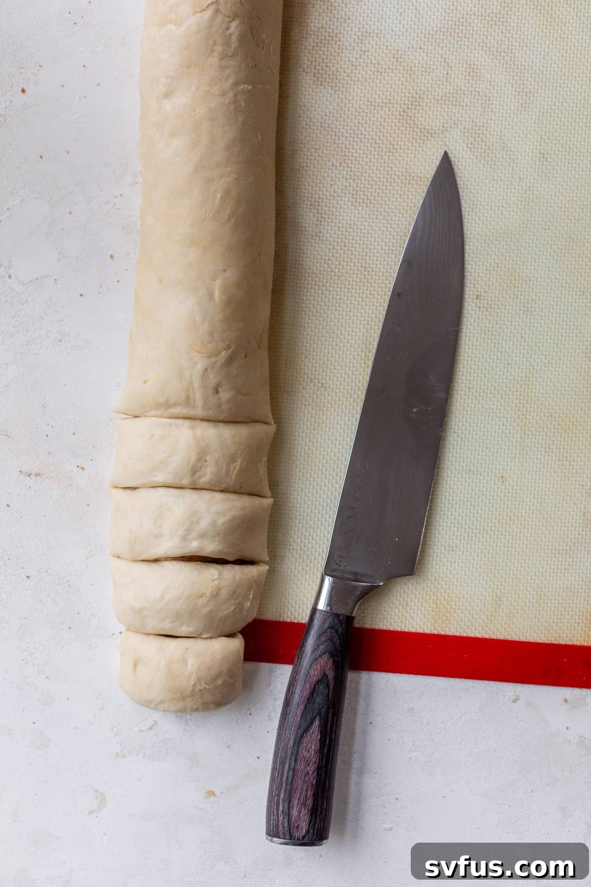 Overhead view of a chef's knife positioned next to a log of vegan cinnamon roll dough on a baking mat, ready for slicing.