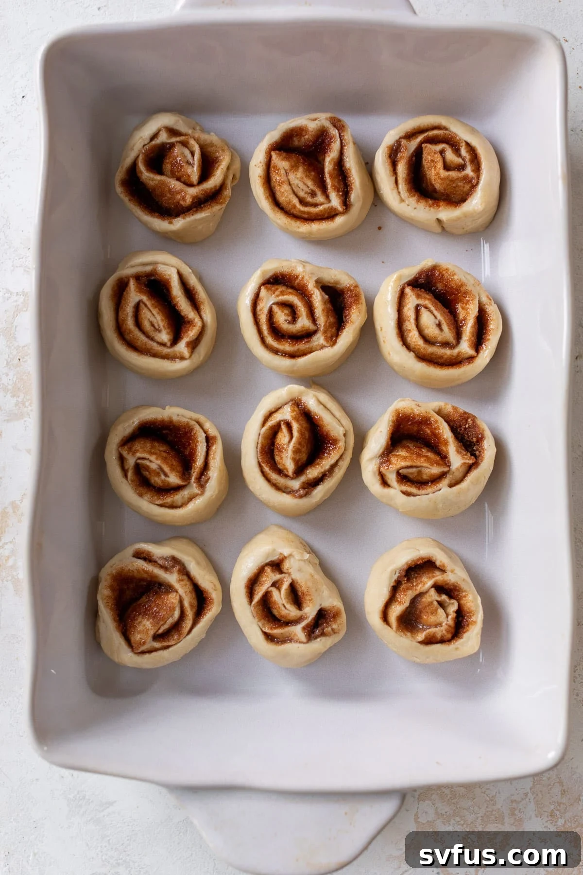 Overhead view of freshly cut, unbaked vegan cinnamon rolls arranged neatly in a white baking dish before their second rise.