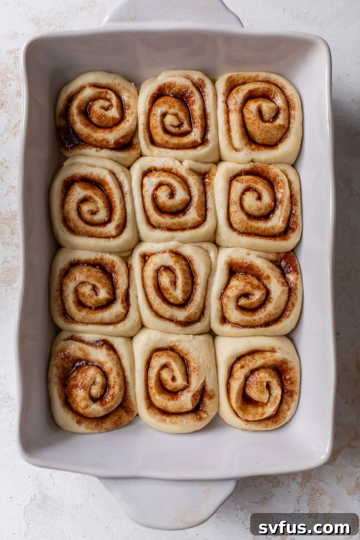 Overhead view of risen and unbaked vegan cinnamon rolls in a white baking dish, showing their increased size.