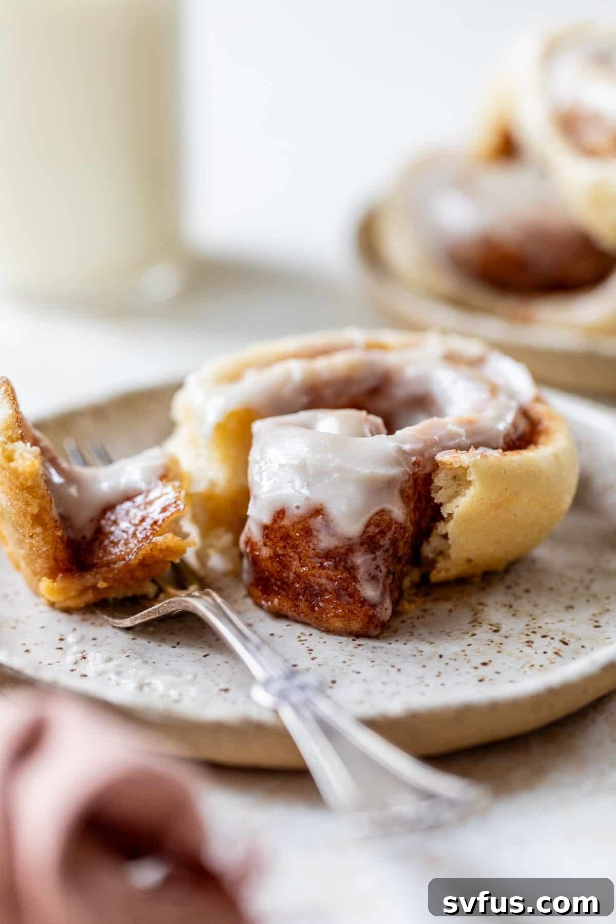 Side view of a fork next to a cream cheese frosting-topped vegan cinnamon roll on a white plate, ready to be eaten.
