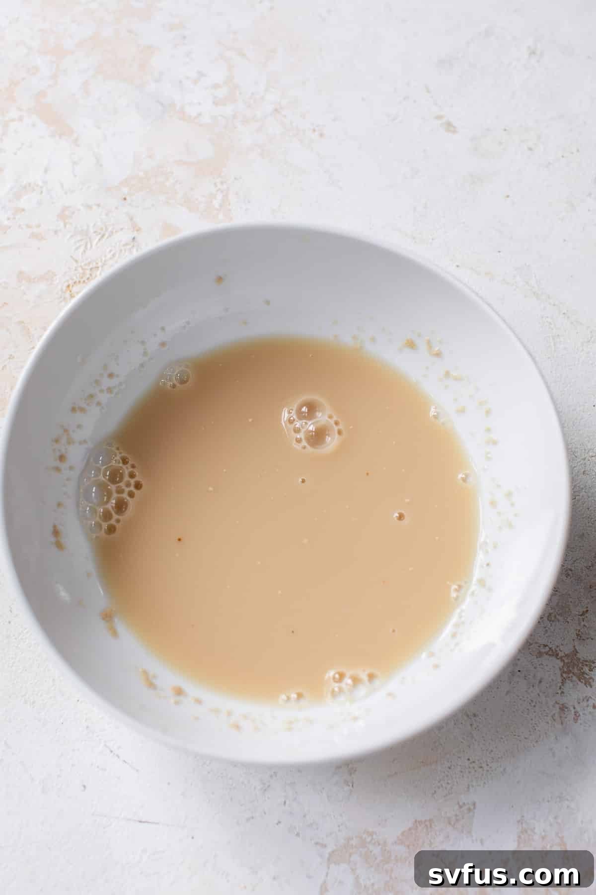 Overhead view of a liquid yeast mixture for vegan cinnamon rolls in a white bowl, showing initial activation.