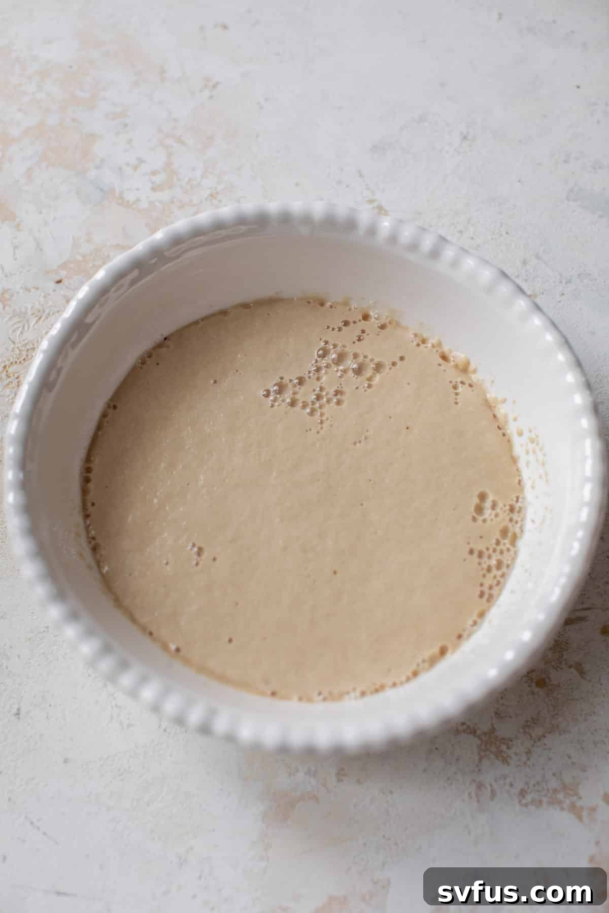 Overhead view of a bubbly and activated yeast mixture for vegan cinnamon rolls in a white bowl, ready for mixing.