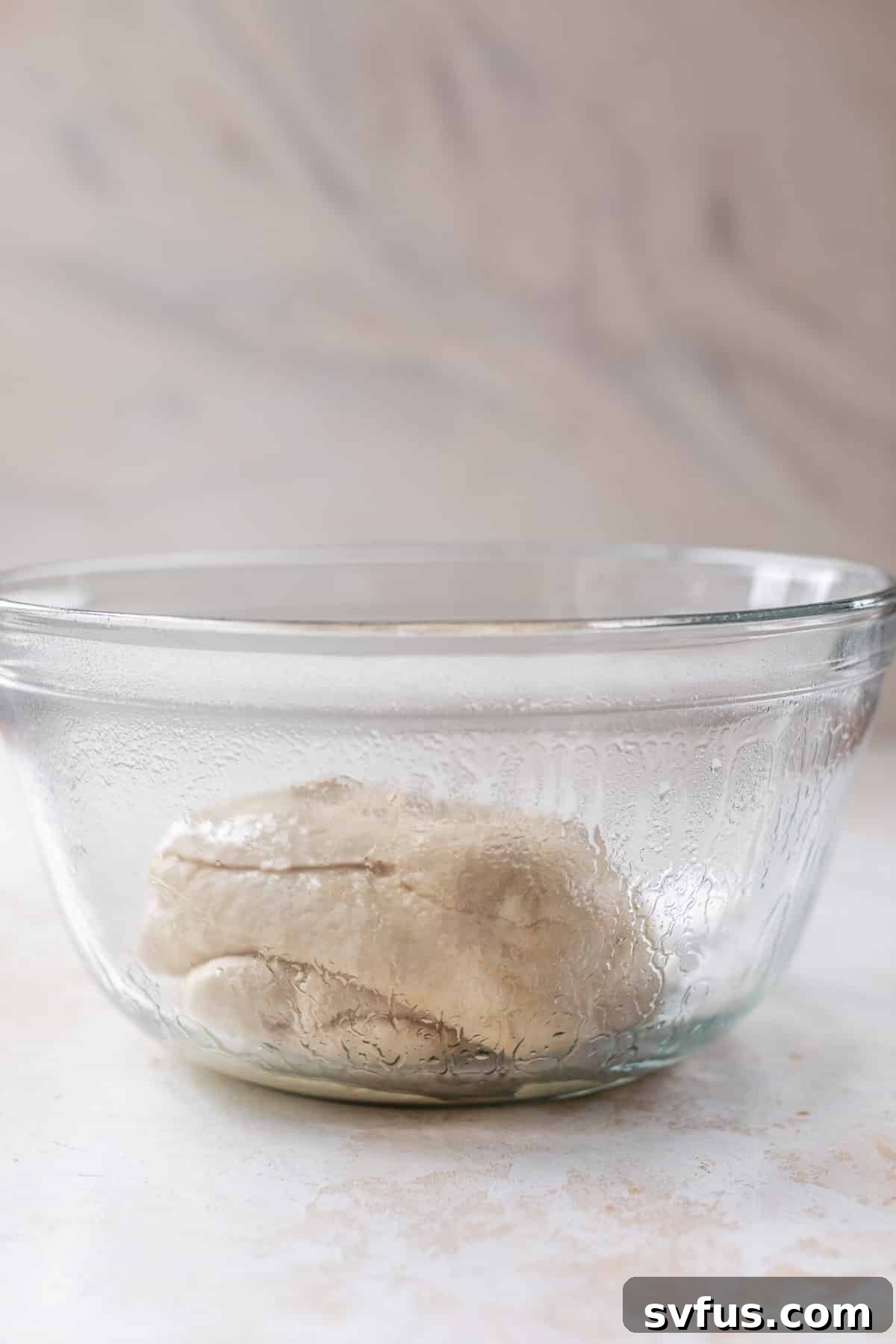 Side view of unrisen vegan cinnamon roll dough in a glass bowl, covered with plastic wrap.