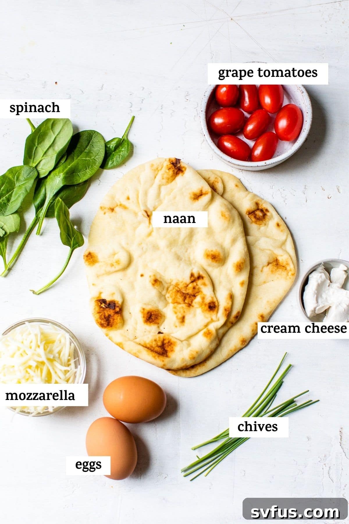 A beautiful flat lay of fresh ingredients including naan bread, a bowl of cream cheese, mozzarella, sliced grape tomatoes, and vibrant spinach, ready to be assembled into delicious breakfast pizzas.