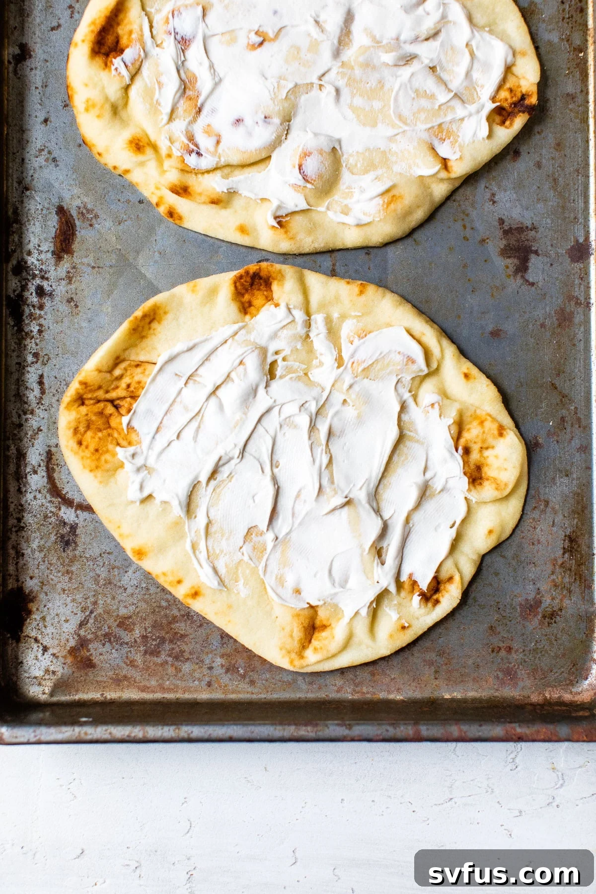 Two pieces of naan bread on parchment paper, each generously topped with a layer of creamy cream cheese, ready for additional toppings.