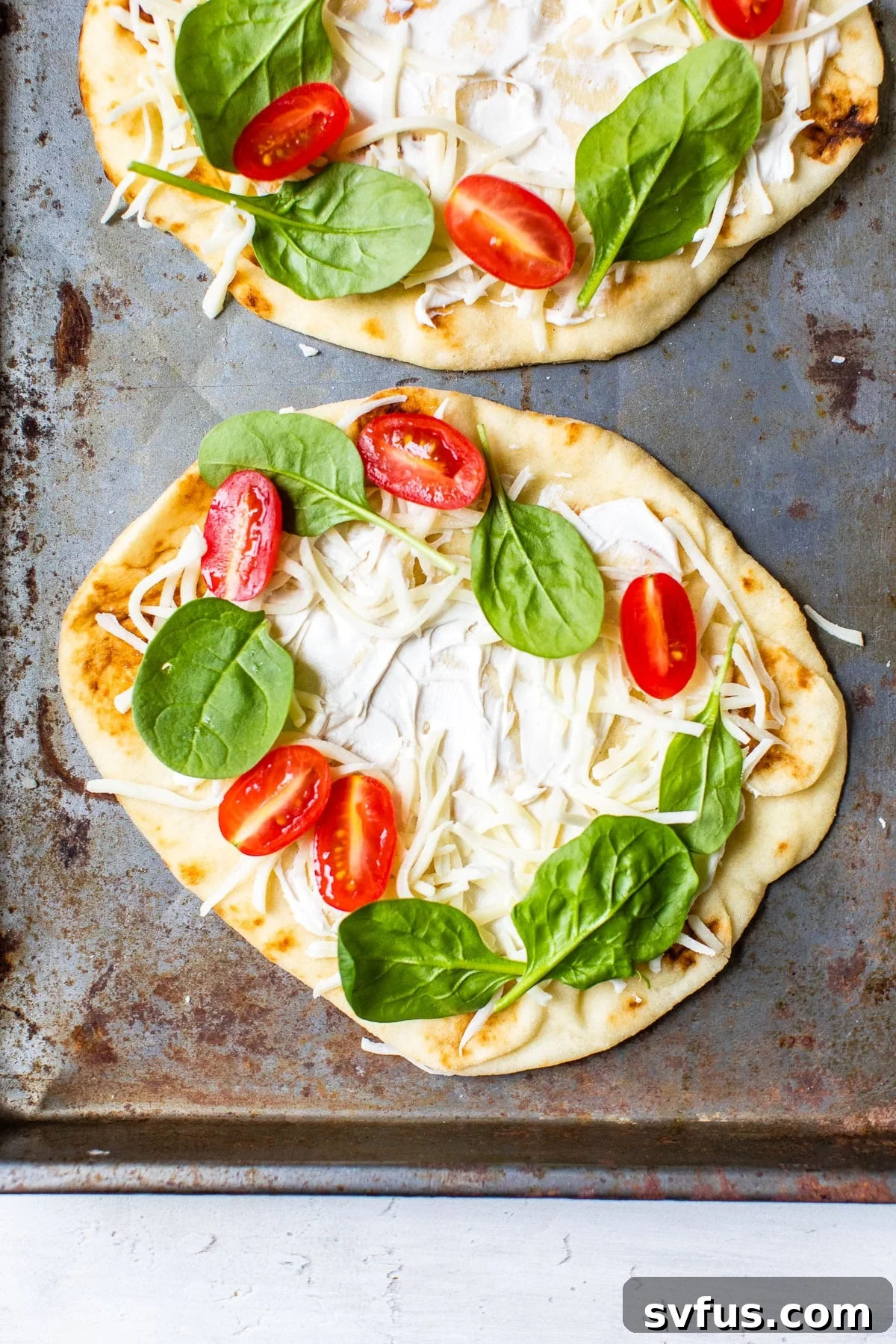 Naan bread pizzas on a baking sheet, topped with melted mozzarella, sliced tomatoes, and fresh spinach, with a perfect well left in the center for an egg.