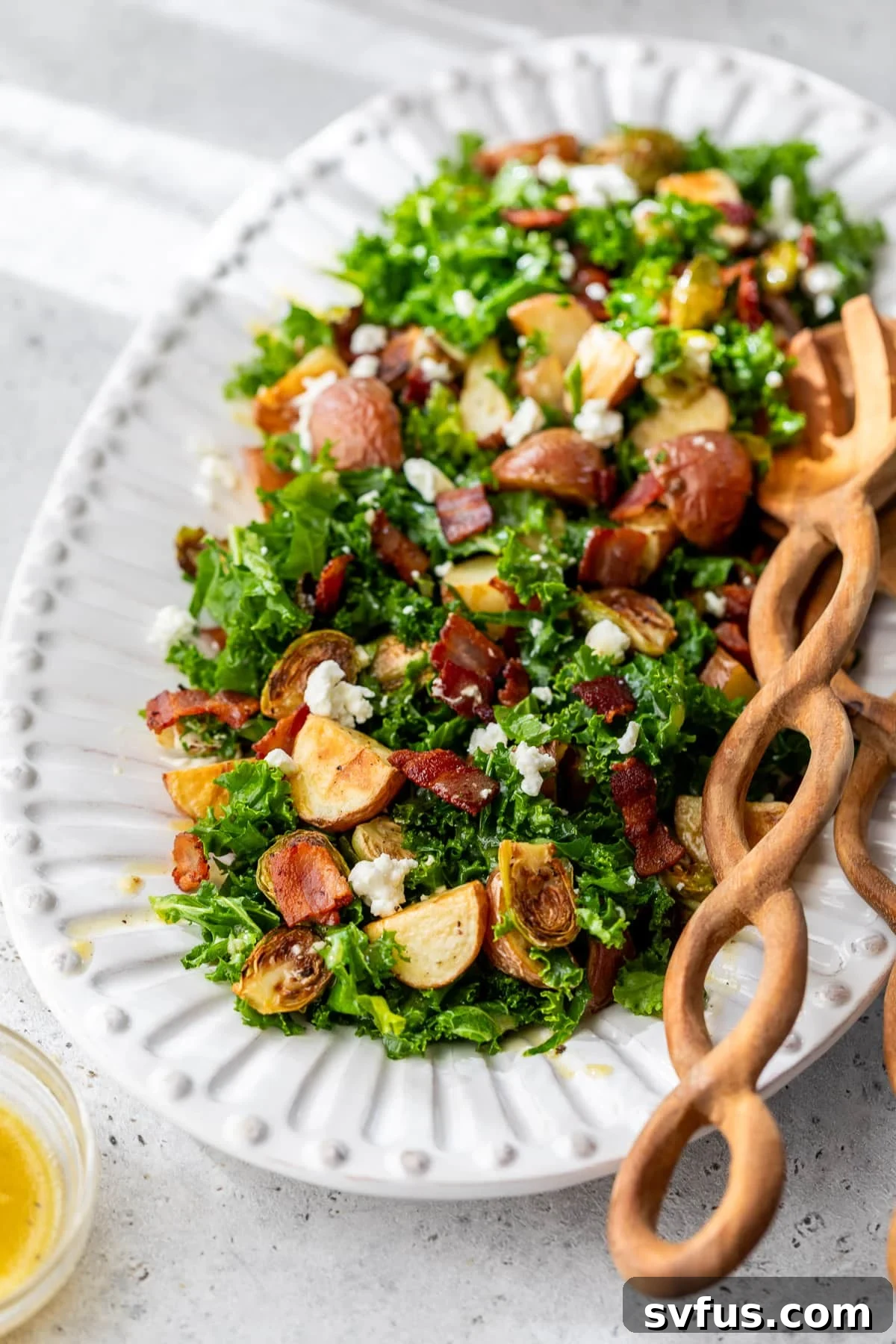 Wooden salad tongs rest beside a large white platter filled with a vibrant Potato Brussels Sprouts Salad, showcasing roasted vegetables, crisp bacon, and creamy goat cheese.