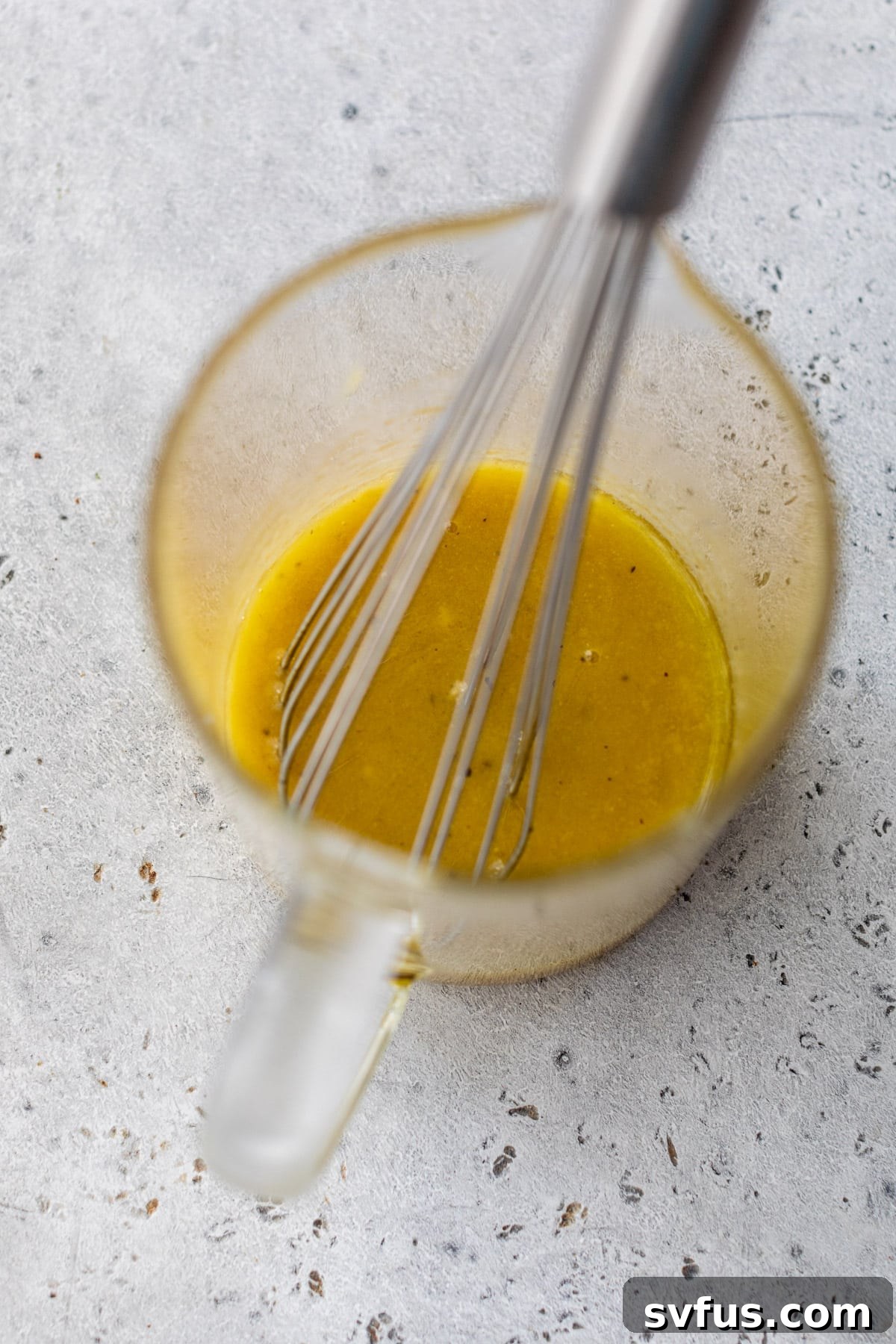A glass measuring cup holds the golden vinaigrette, being whisked to combine the ingredients into a smooth dressing.
