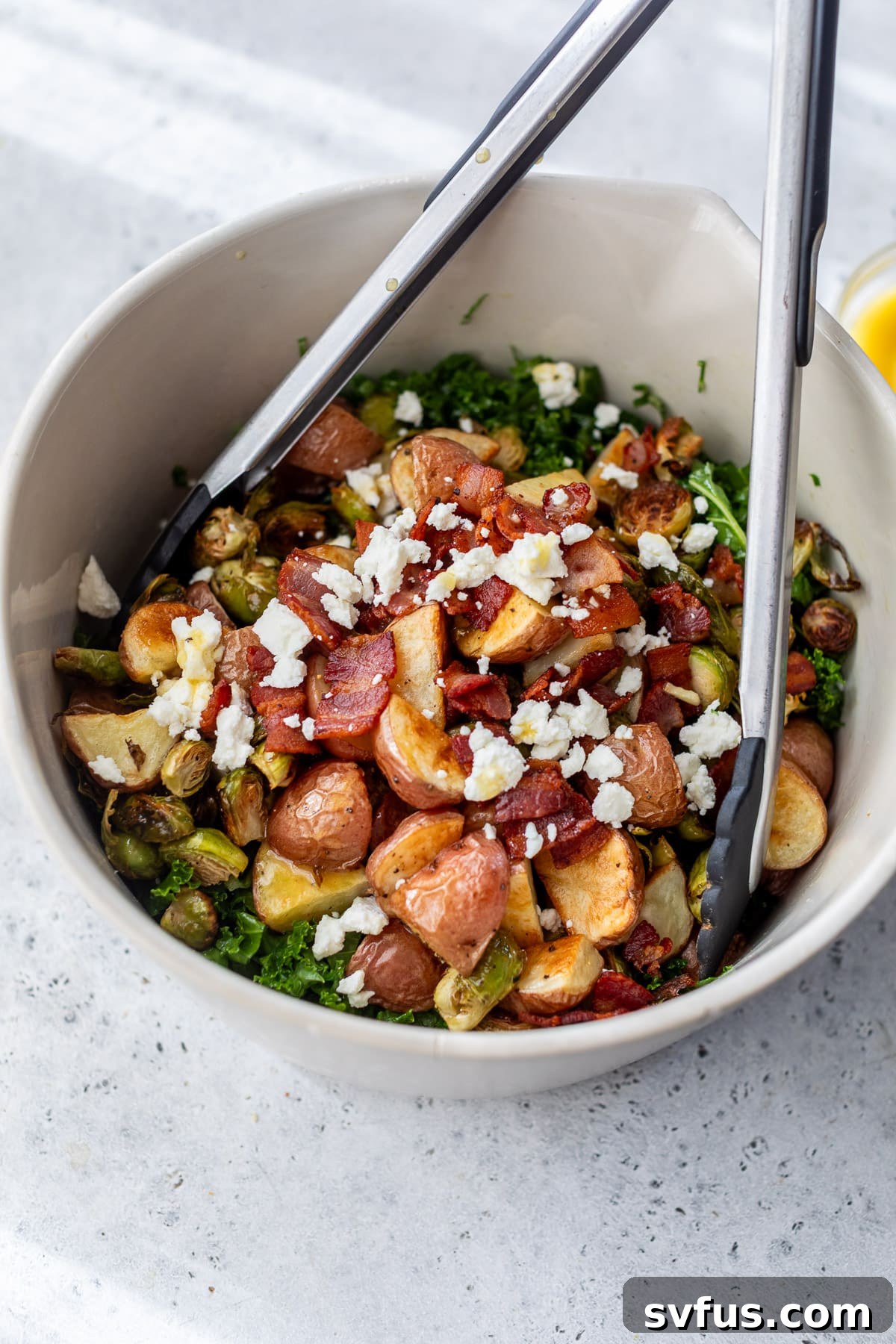 Salad tongs are actively mixing the Potato Brussels Sprouts Salad in a large white bowl, ensuring all ingredients are evenly coated with dressing.