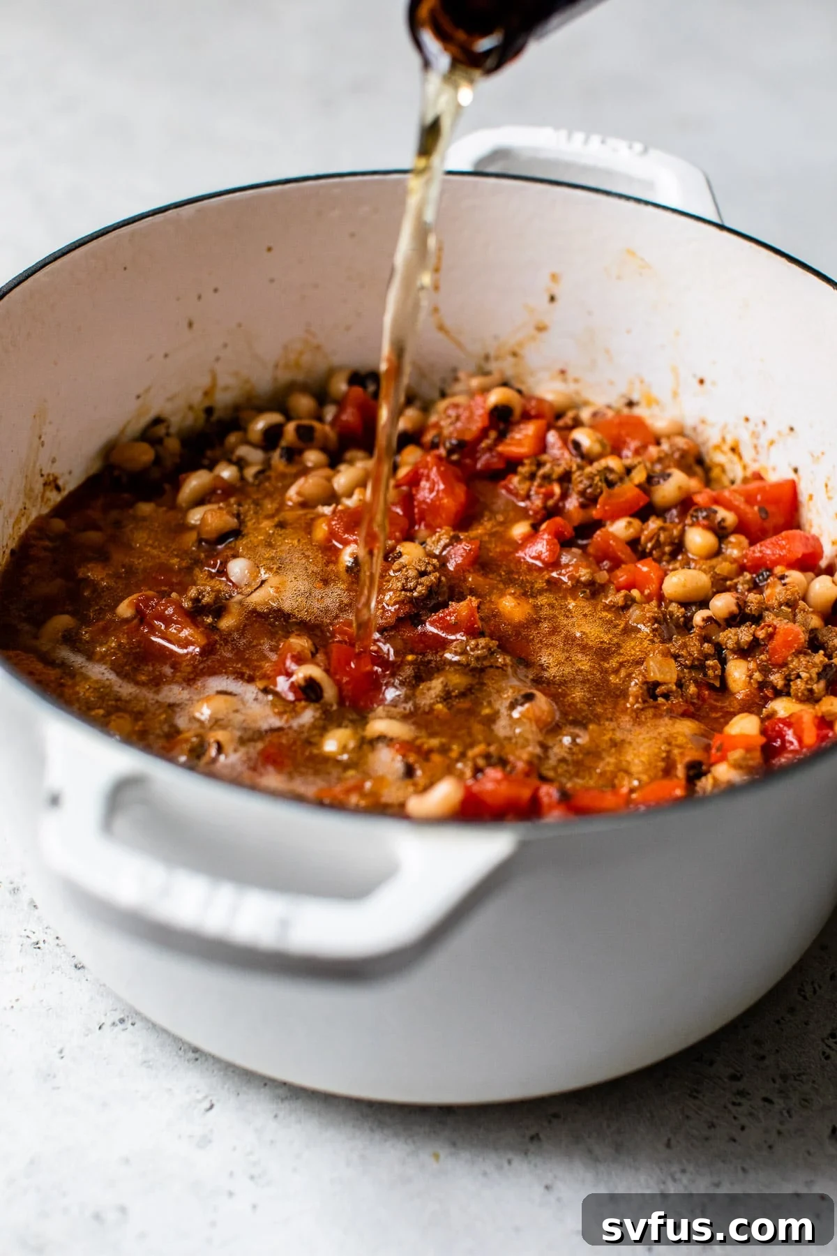 A hand pouring a bottle of beer into the simmering Black Eyed Peas Beer Chili in a Dutch oven, adding the final liquid component.