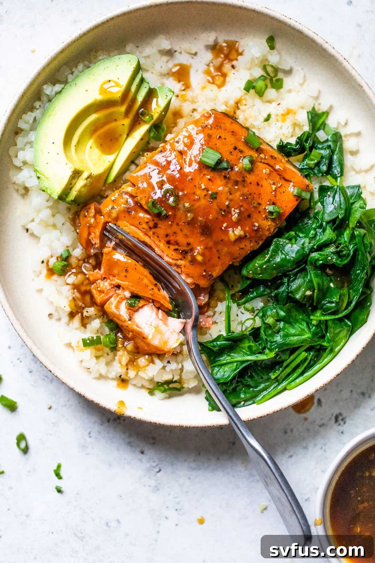 A beautifully arranged bowl filled with golden-brown teriyaki glazed salmon, fluffy cauliflower rice, vibrant cooked spinach, and sliced avocado