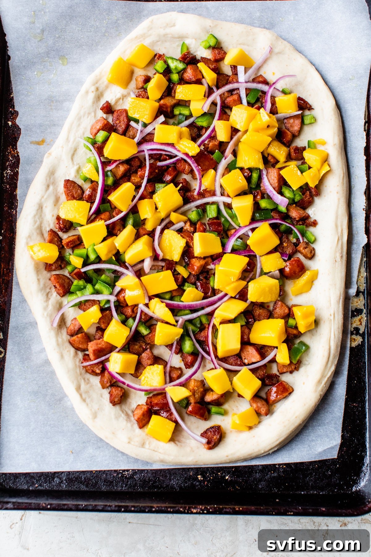 An uncooked pizza on a baking sheet, showcasing a colorful array of chorizo, fresh mango, vibrant green poblano pepper, and thinly sliced red onion, ready for the oven.