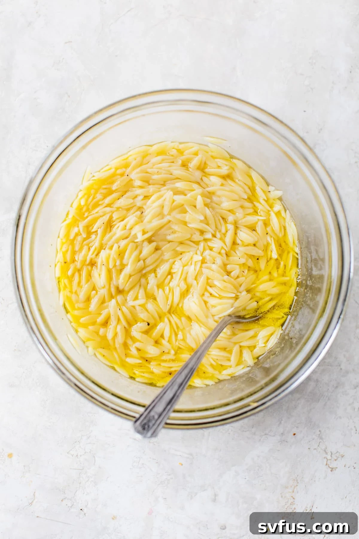 A glass bowl filled with cooked orzo pasta soaking in a vibrant lemon and olive oil dressing.