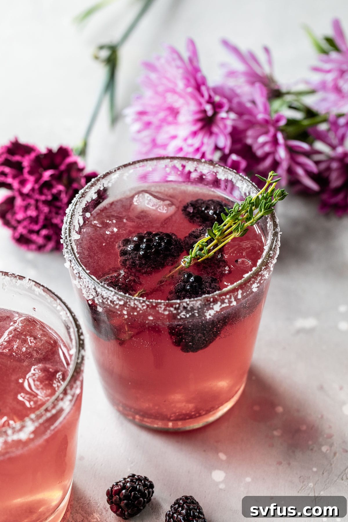purple cocktail with blackberries and purple flowers in the background, garnished with a thyme sprig