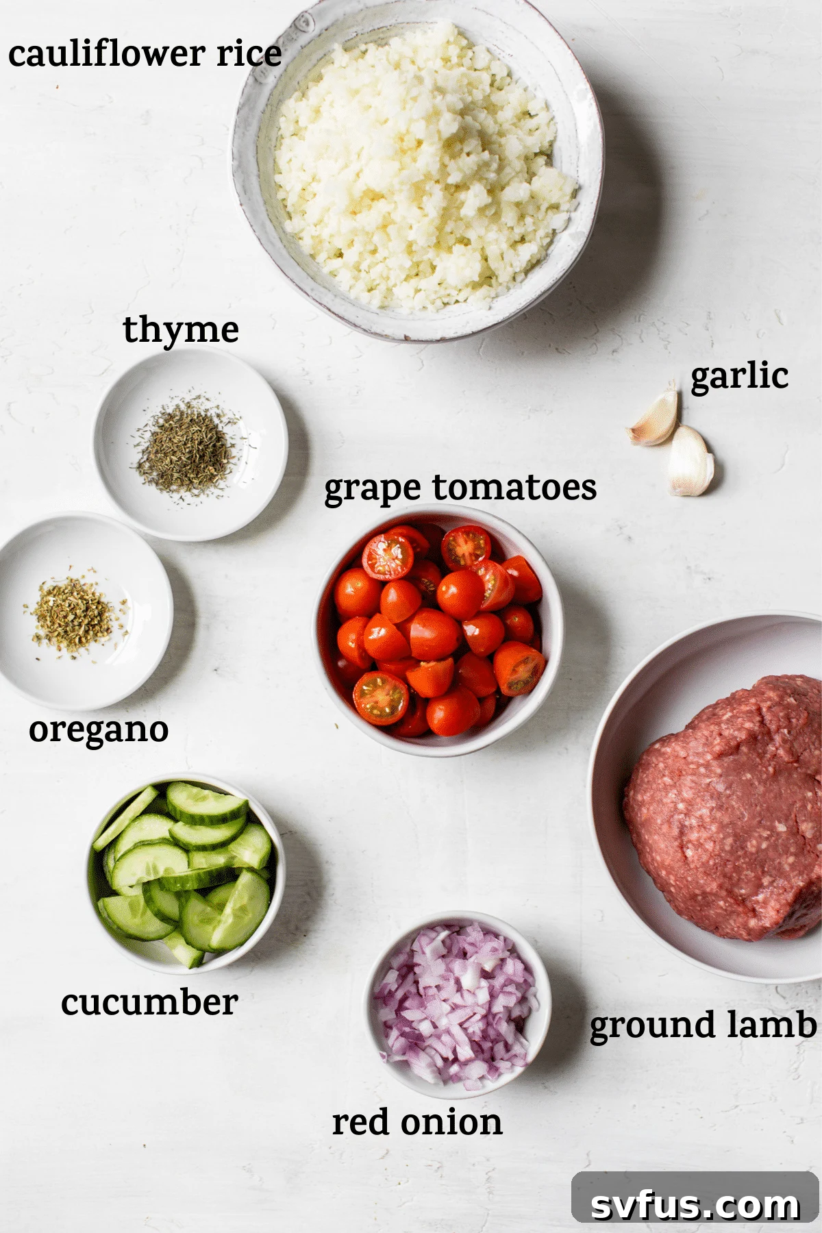 A flat lay of fresh ingredients laid out on a white surface, including raw ground lamb, a bag of frozen cauliflower rice, red onions, cucumbers, grape tomatoes, and a small bowl of herbs, ready for the Mediterranean Lamb Bowl recipe.