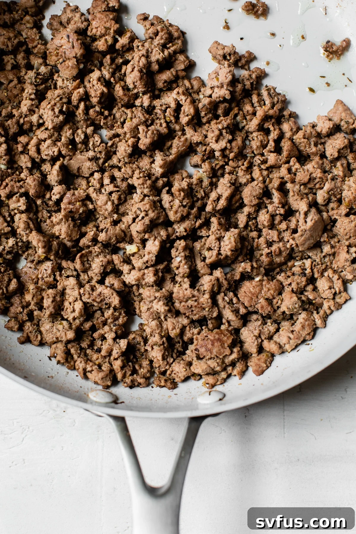 Sizzling ground lamb seasoned with garlic, thyme, and oregano, cooking in a large non-stick skillet over medium heat, releasing rich aromas.