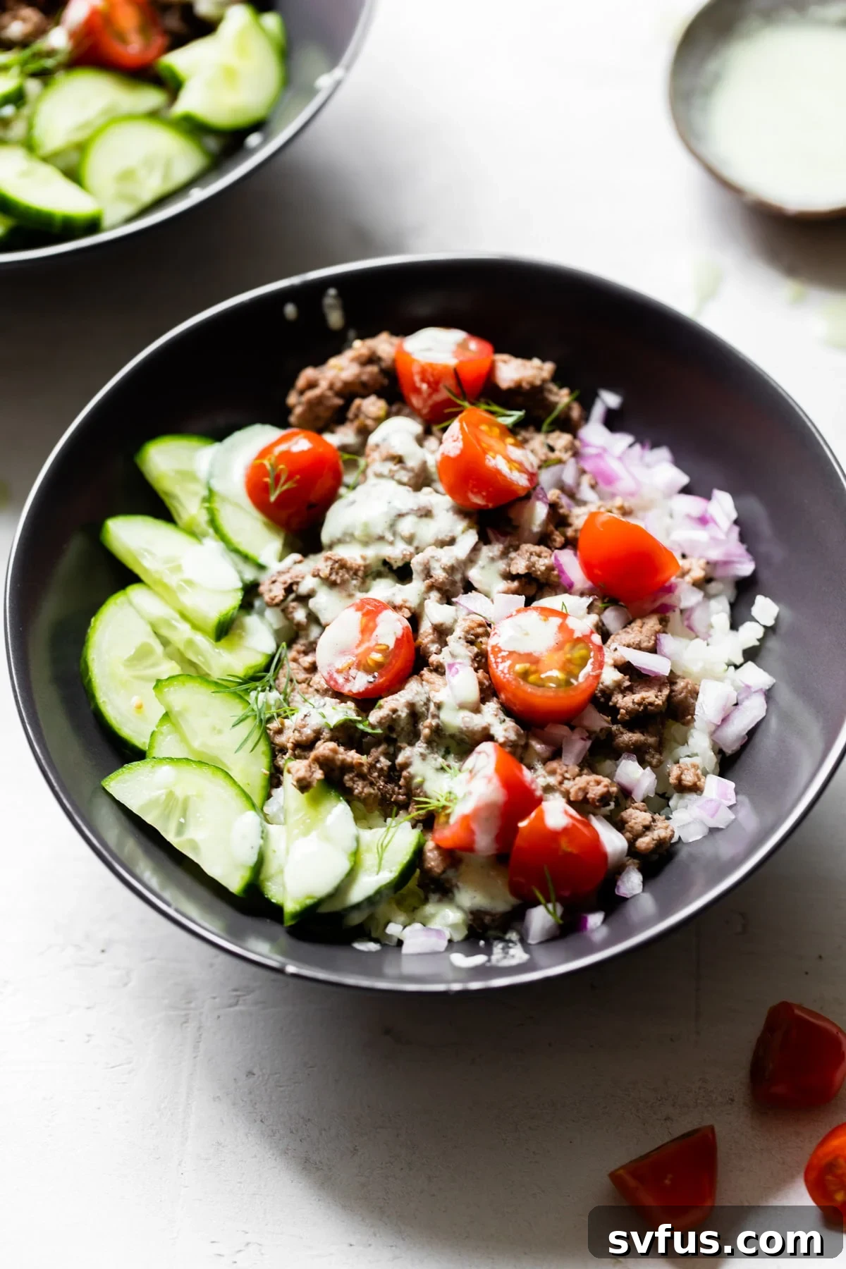 A beautifully assembled Mediterranean Lamb Bowl, showcasing a bed of cauliflower rice topped with savory lamb, vibrant cucumber slices, juicy cherry tomatoes, and a sprinkle of fresh red onion, all generously drizzled with creamy tzatziki.