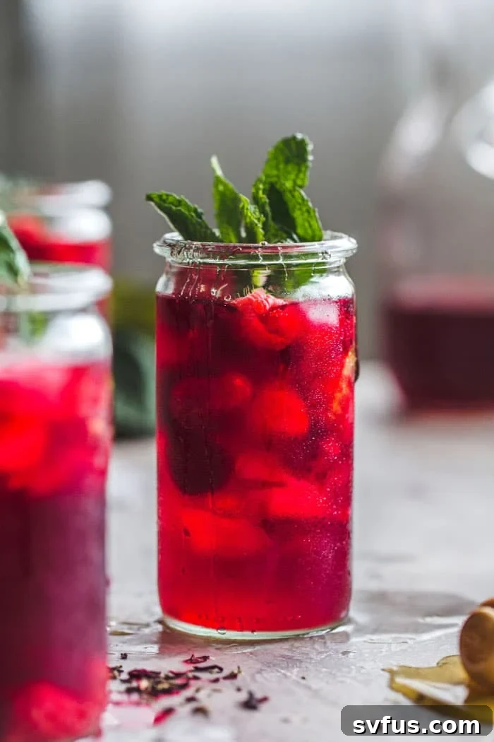 A tall glass of Raspberry Hibiscus Iced Tea garnished with fresh mint and raspberries, on a wooden surface.