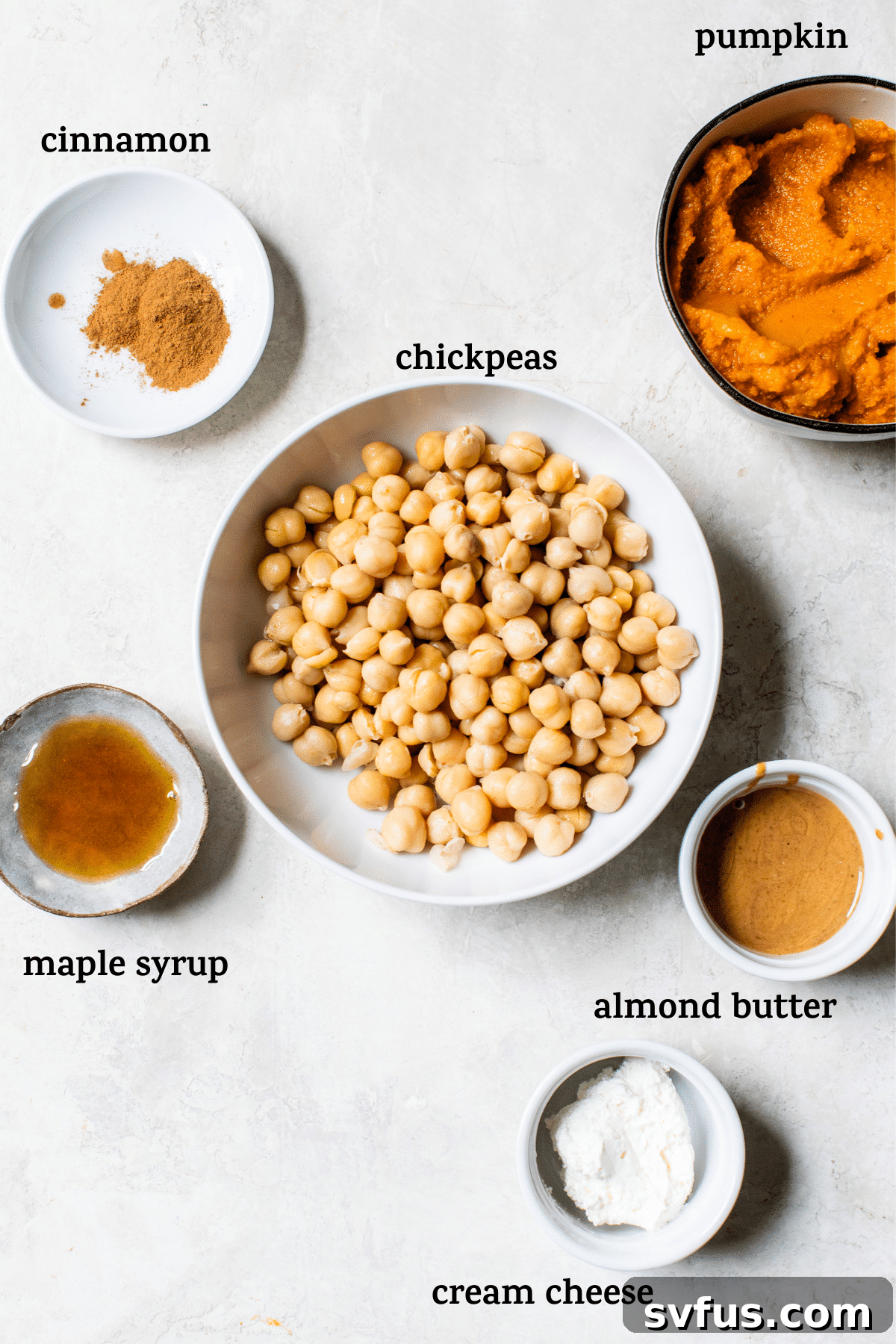 Various ingredients for pumpkin hummus laid out in small bowls on a wooden table, including chickpeas, pumpkin puree, cream cheese, almond butter, maple syrup, and cinnamon.