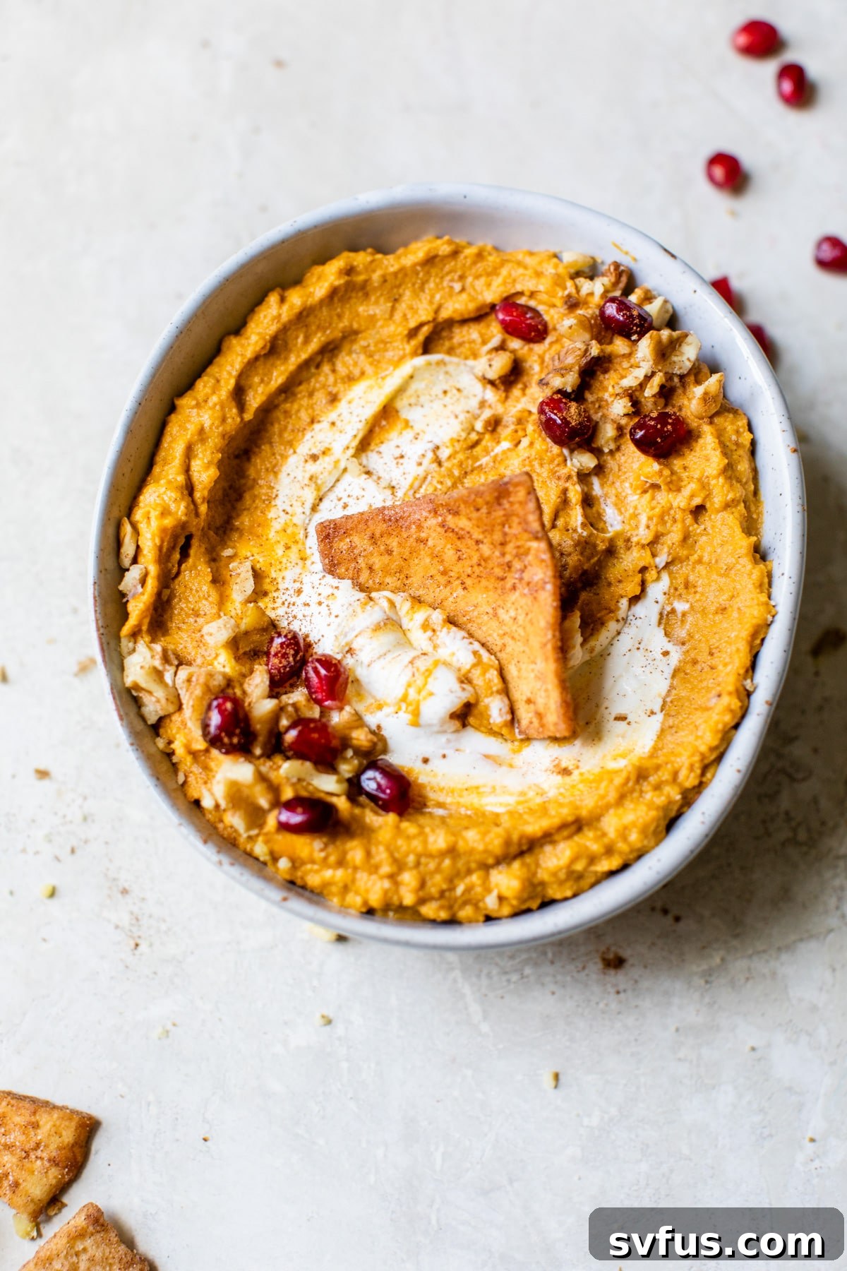 A bowl of pumpkin hummus garnished with nuts and a swirl of cream cheese, surrounded by cinnamon sugar pita chips on a rustic wooden surface.