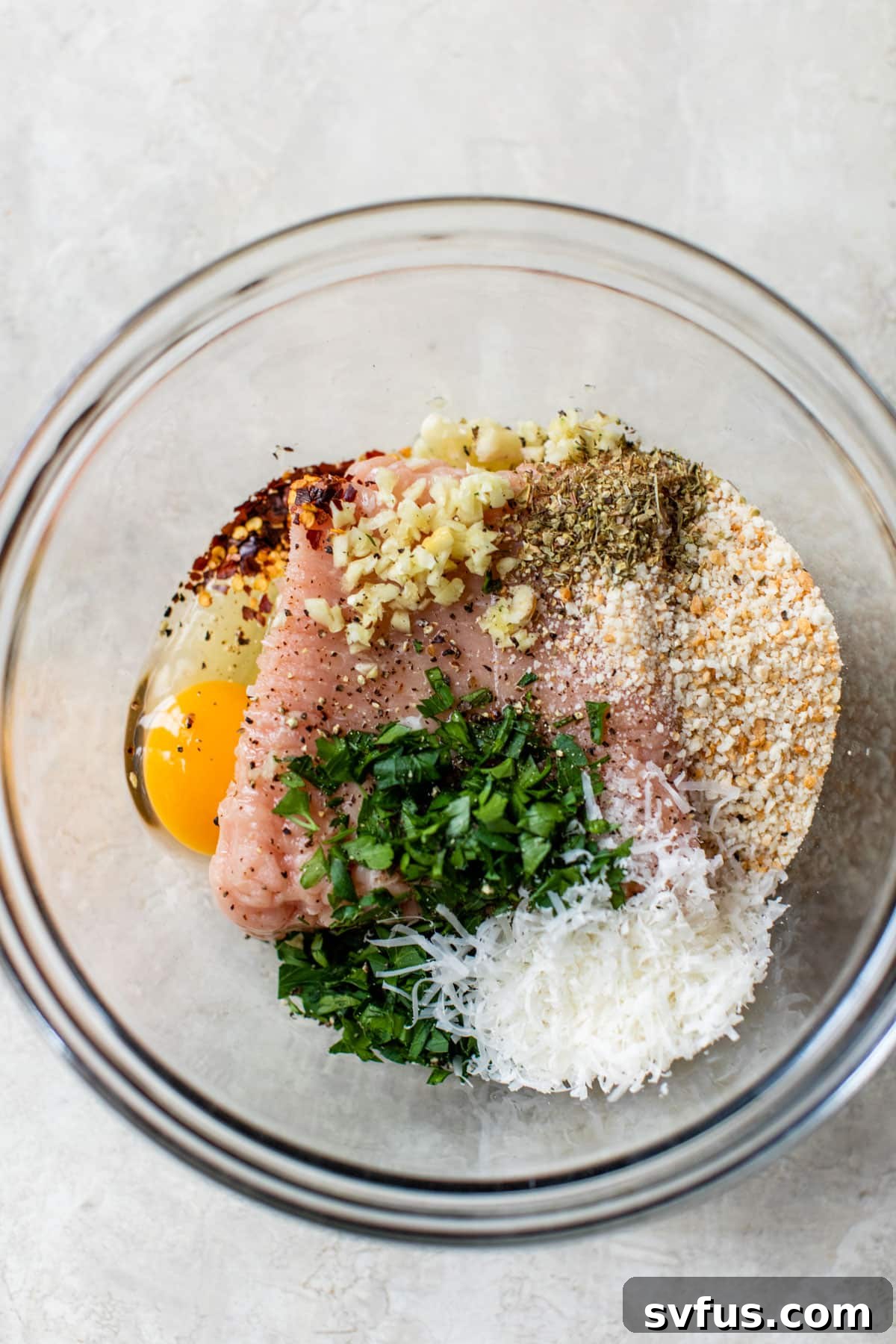 A large glass bowl containing all the raw ingredients for chicken meatballs: ground chicken, egg, chopped parsley, breadcrumbs, grated parmesan, and minced garlic, before mixing.
