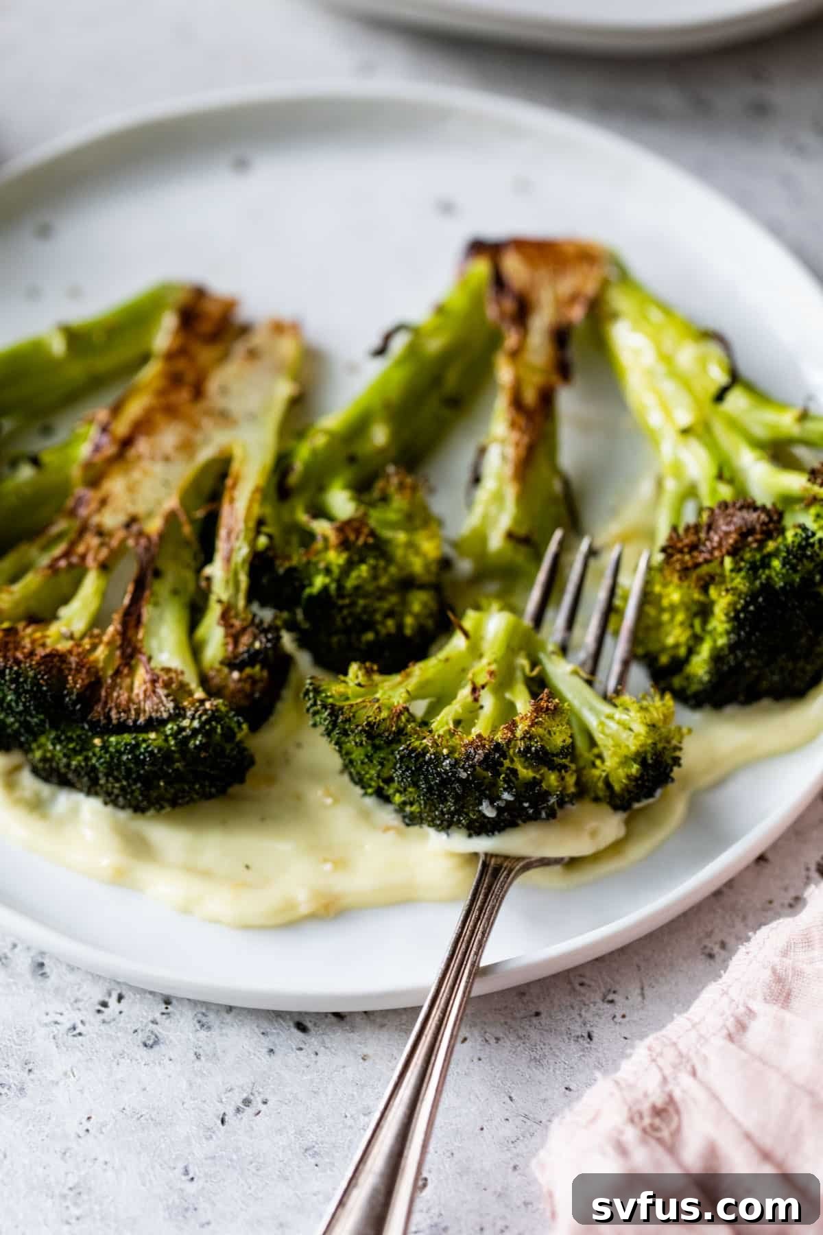 A hand gently cutting into a roasted broccoli crown on a plate with a fork, emphasizing its tender texture.