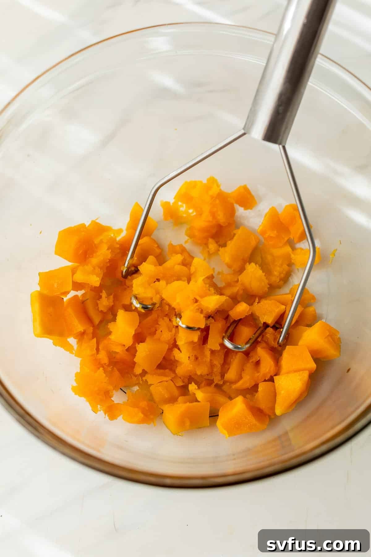 mashing softened butternut squash cubes in a glass bowl with a potato masher, preparing it for the bread batter.
