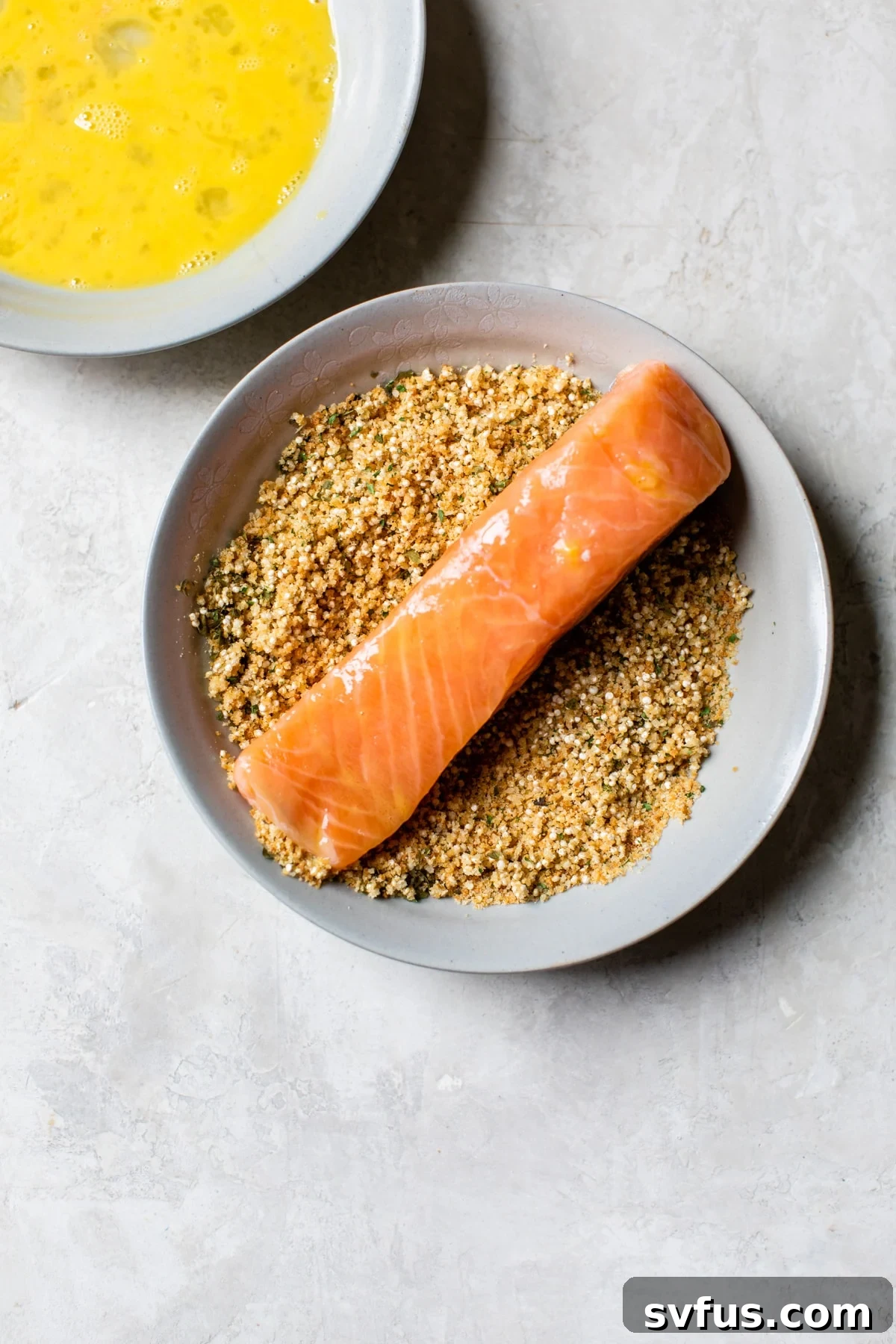 A salmon fillet being coated with the quinoa and breadcrumb mixture in a bowl, showing a thick and even layer of crust.