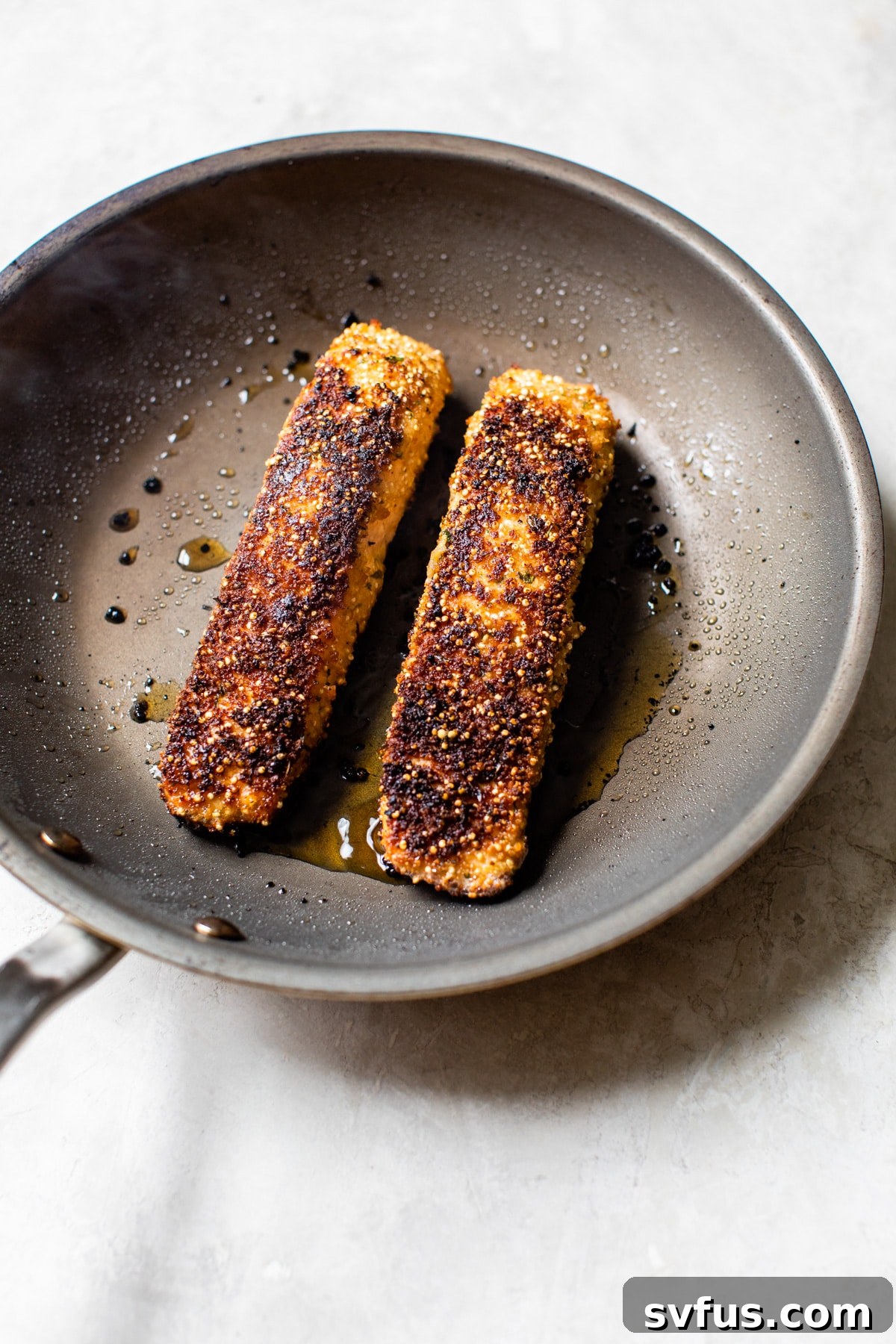 Two Quinoa Crusted Salmon fillets cooking in a nonstick skillet with a lid on, showing the steam and browning process.
