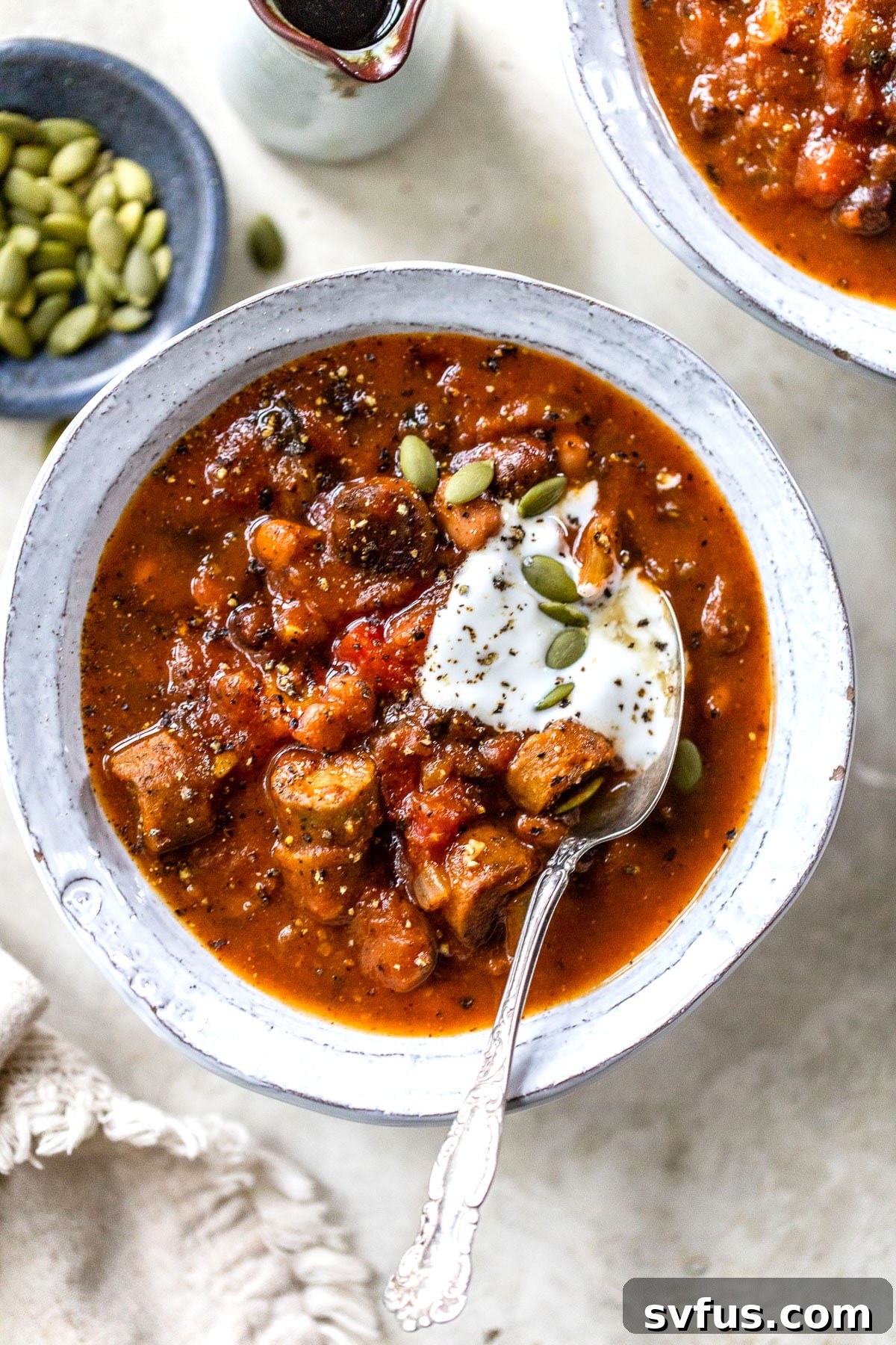 a bowl of chili with a spoon in it beside a small dish of pumpkin seeds