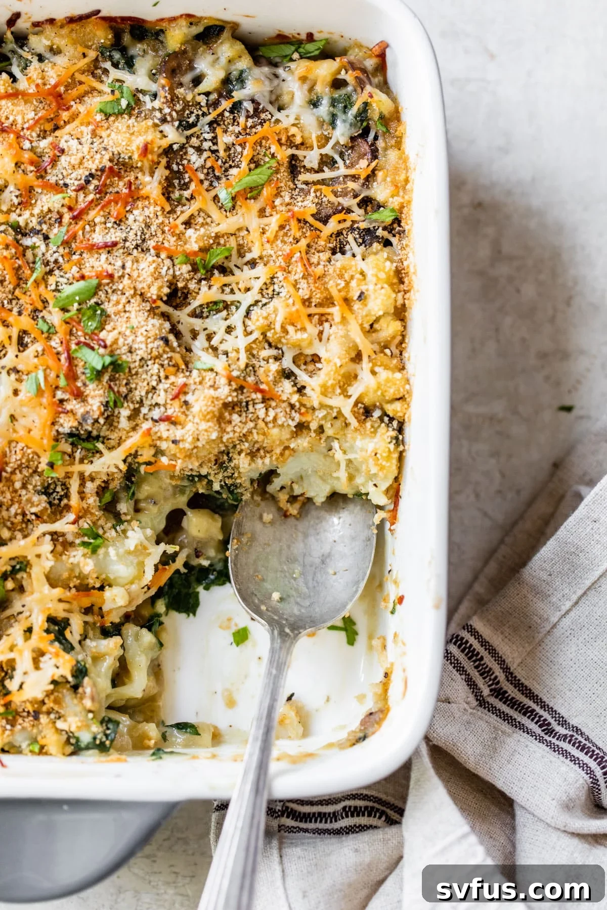 A golden brown vegan cauliflower gratin in a casserole dish, topped with crispy breadcrumbs and a spoon ready to serve.