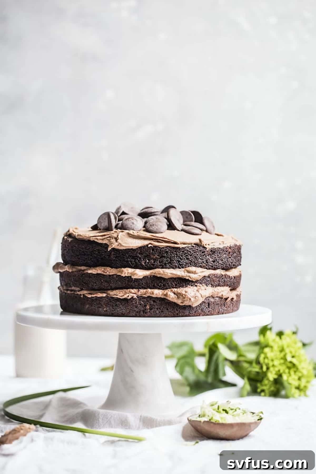 Tall, three-layer Vegan Chocolate Zucchini Cake on a cake stand, frosted with dairy-free chocolate buttercream and garnished with chocolate shavings.