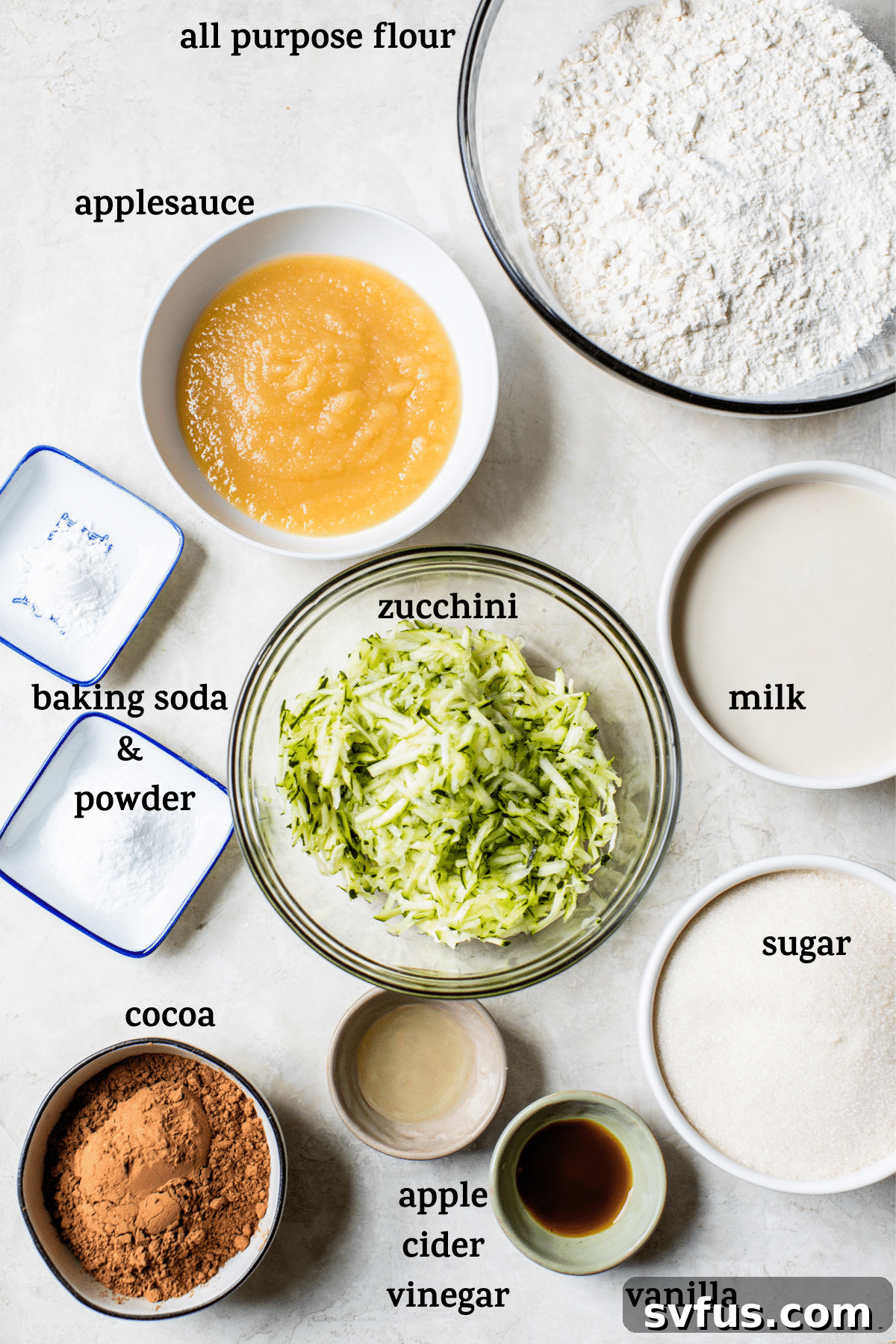 Various vegan chocolate zucchini cake ingredients laid out on a white surface, including flour, cocoa, sugar, zucchini, applesauce, and plant-based milk.