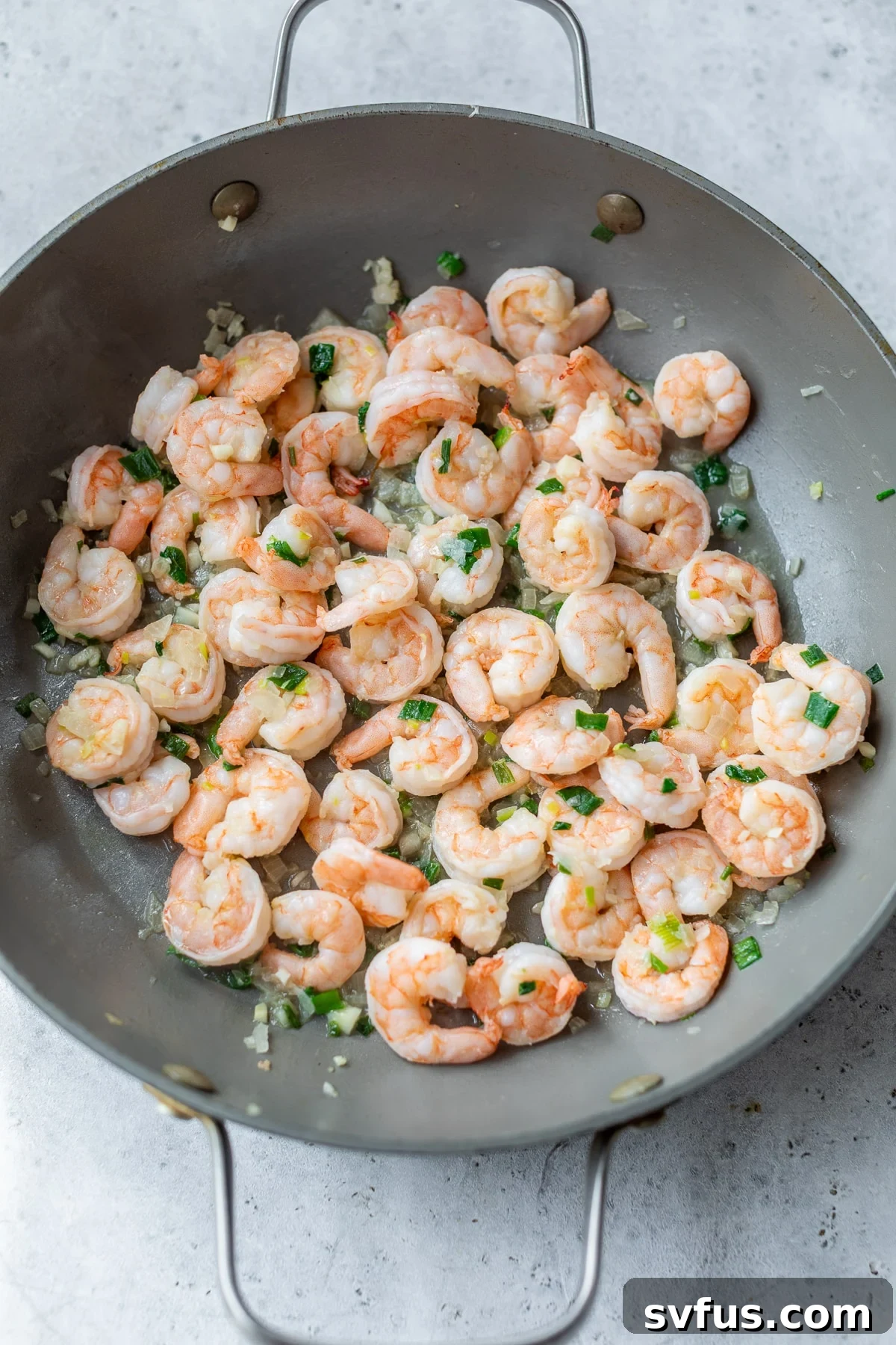 Shrimp cooking in a large skillet alongside aromatics, turning pink and ready for the next step.